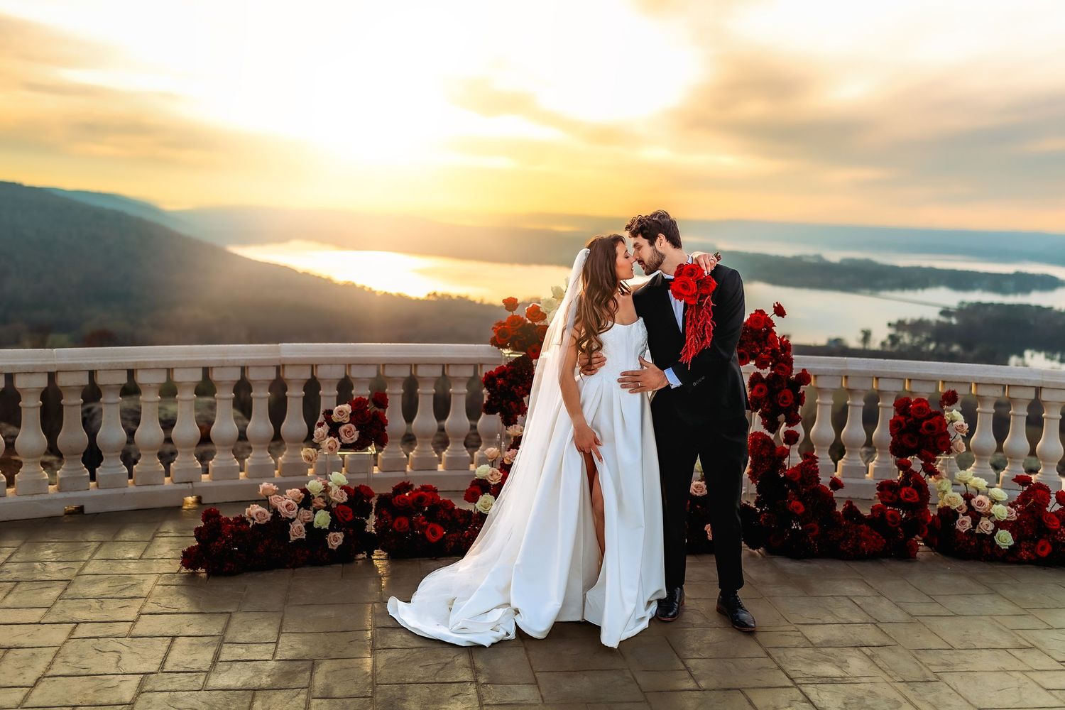 Bride and groom share romantic sunset kiss on elegant balcony adorned with red and white floral arrangements overlooking mountains.