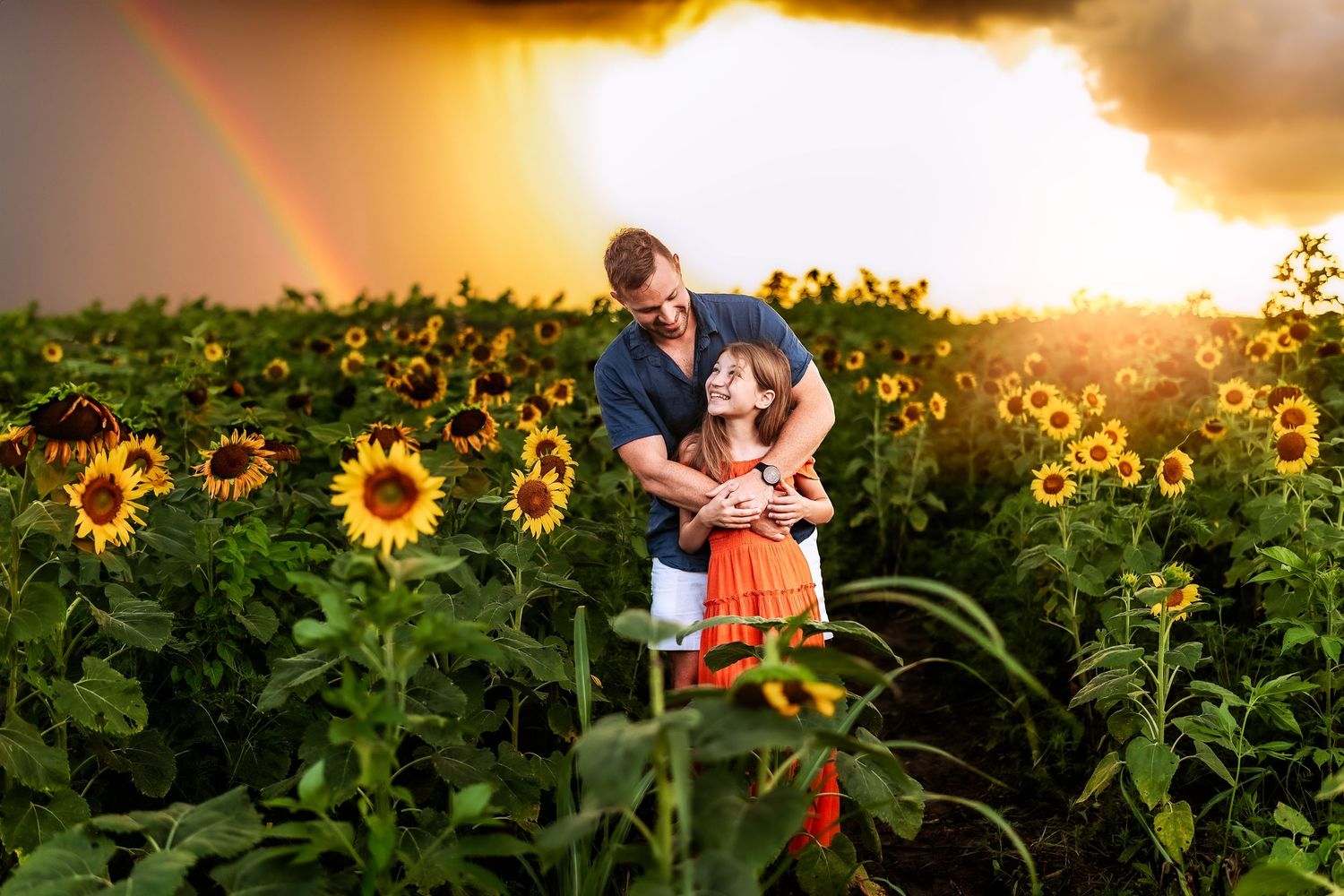 Couple embracing in a sunflower field at golden hour with a rainbow in the stormy sky behind them.