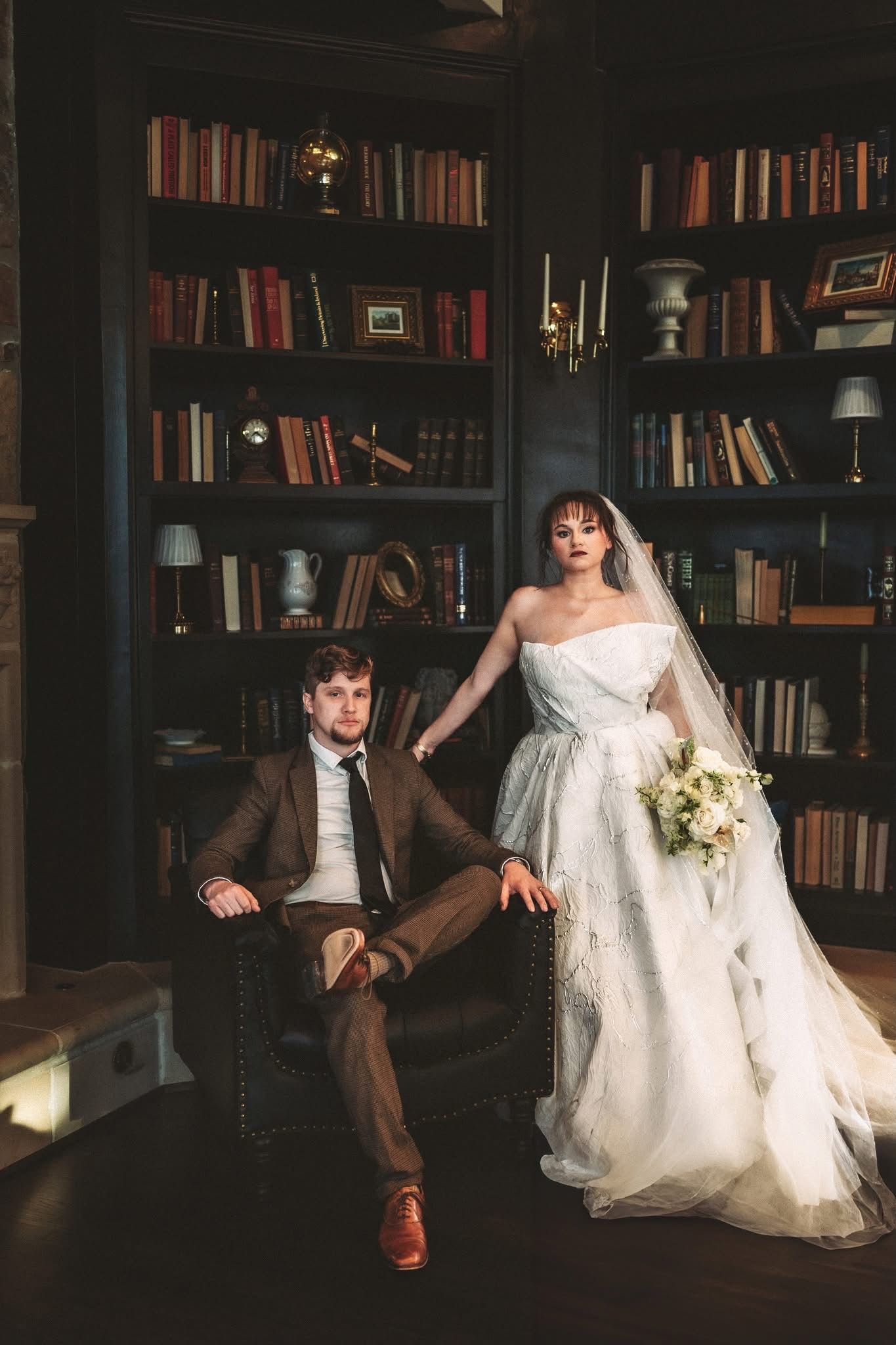 Bride in white gown holding bouquet stands beside groom in brown suit seated in leather chair before dark bookshelf library.