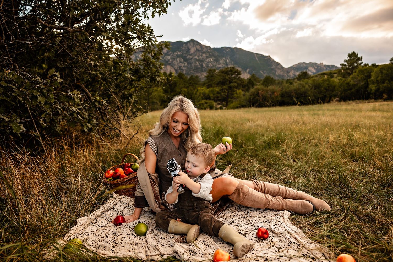 Smiling blonde mother and toddler sit on a cream blanket with apples in a basket in a grassy field with mountains behind at sunset.