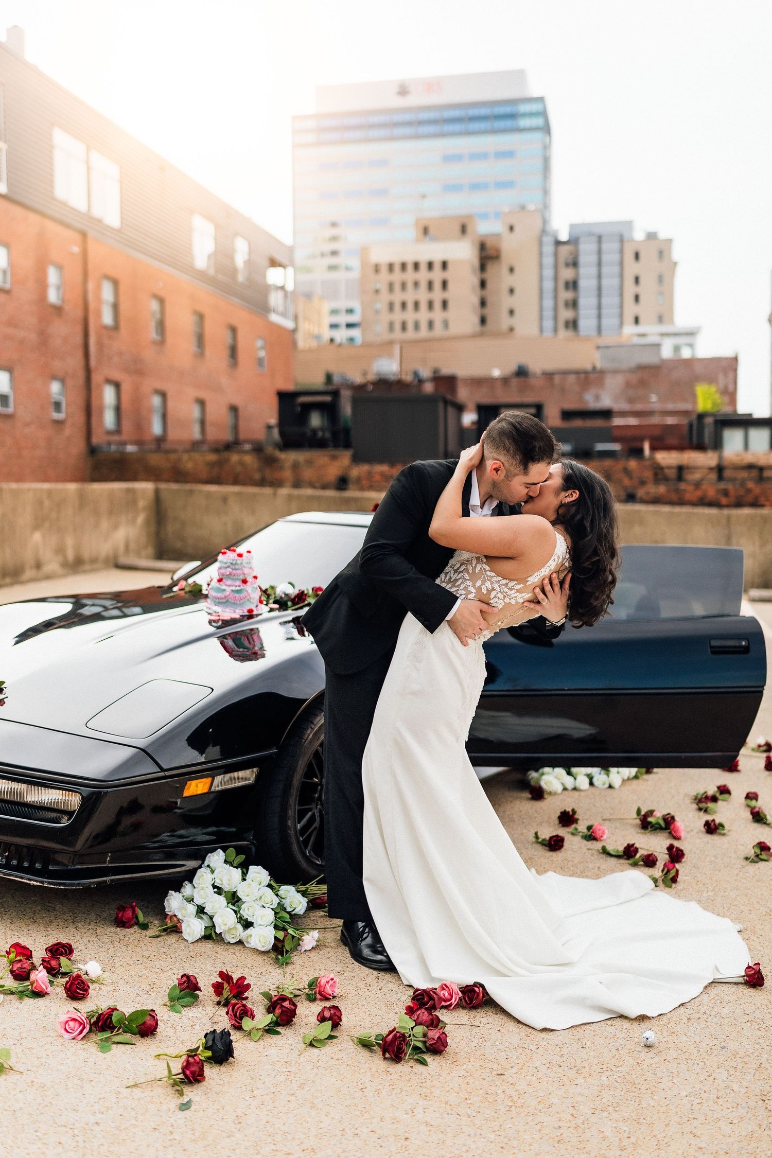 Bride and groom share a romantic dip kiss beside a black Corvette adorned with roses on a rooftop with city skyline backdrop.