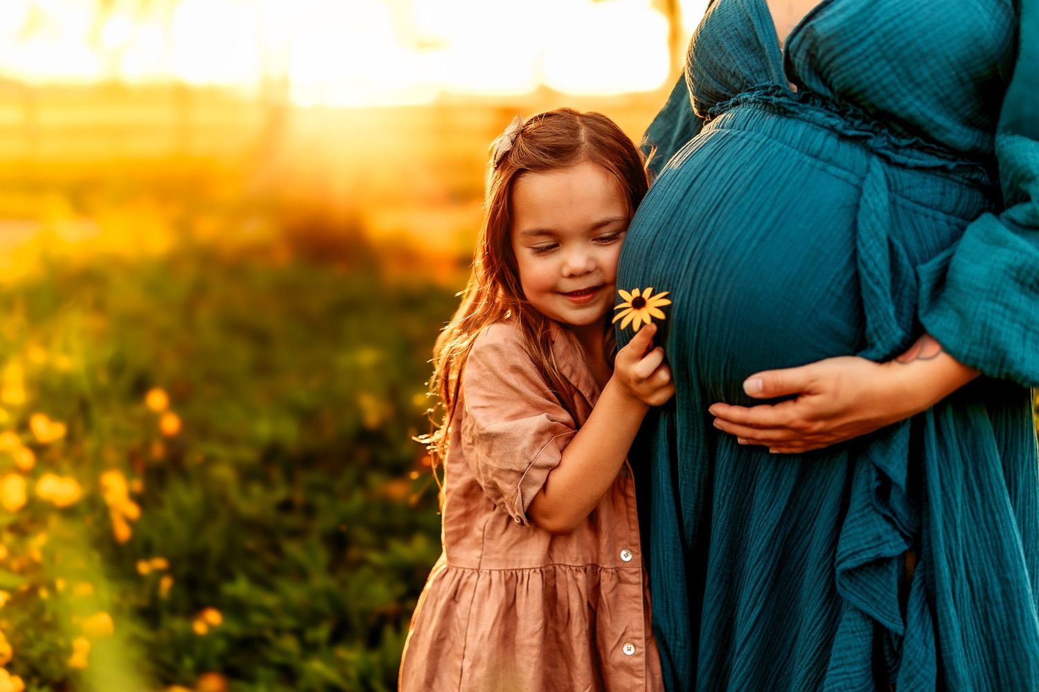 A little girl in a pink dress holds a yellow flower against a pregnant woman's belly at golden hour.