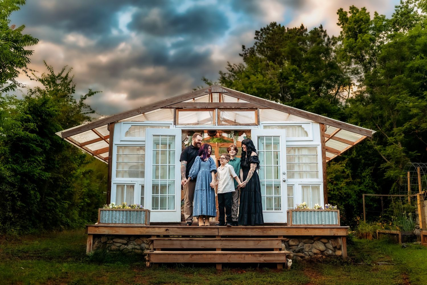 Family posing on wooden deck of charming greenhouse cottage with large windows under dramatic cloudy sky.