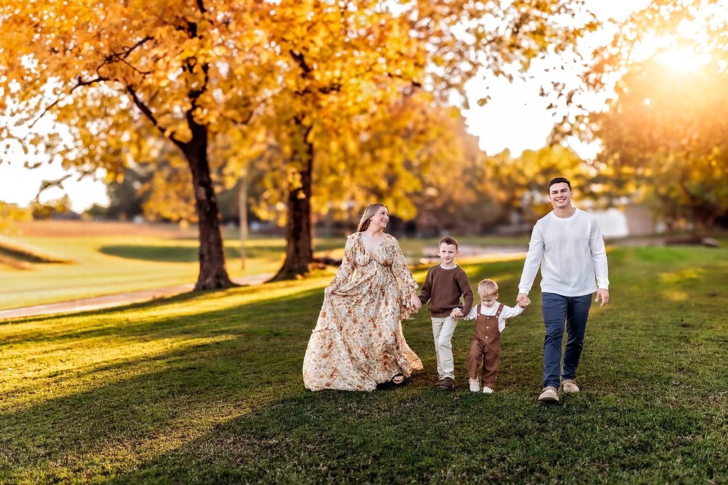 Family of four walking hand-in-hand on green grass beneath golden autumn trees during a warm sunset.