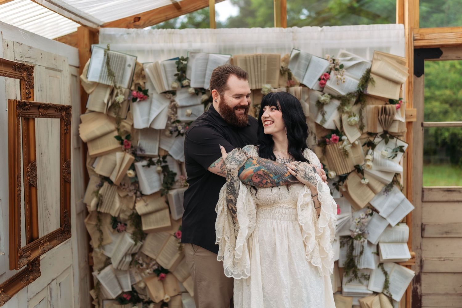Tattooed couple smiling together at rustic wedding venue with decorative book page backdrop and floral accents.