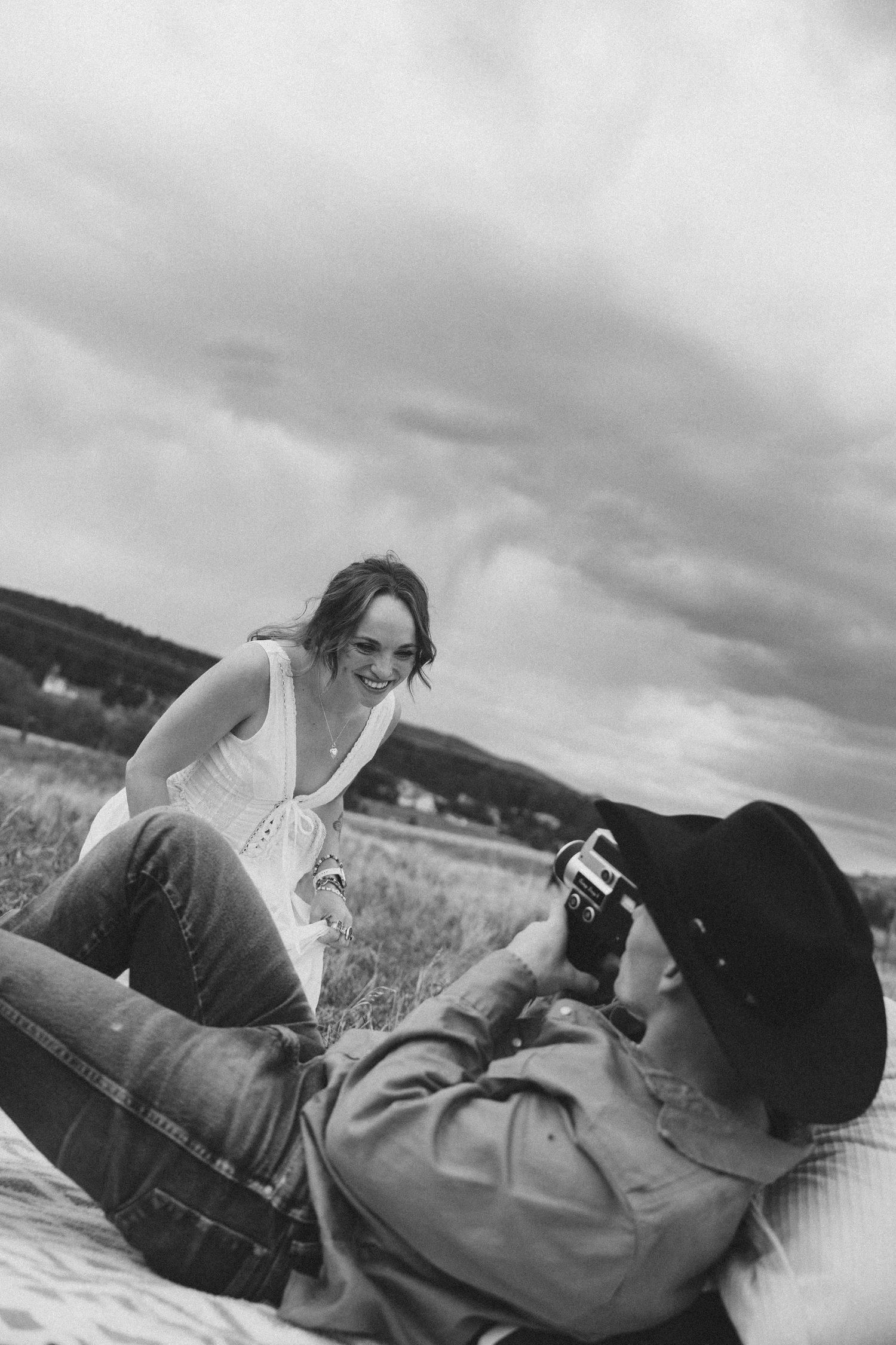 Black and white photo of a man in a cowboy hat photographing a smiling woman outdoors under a cloudy sky.