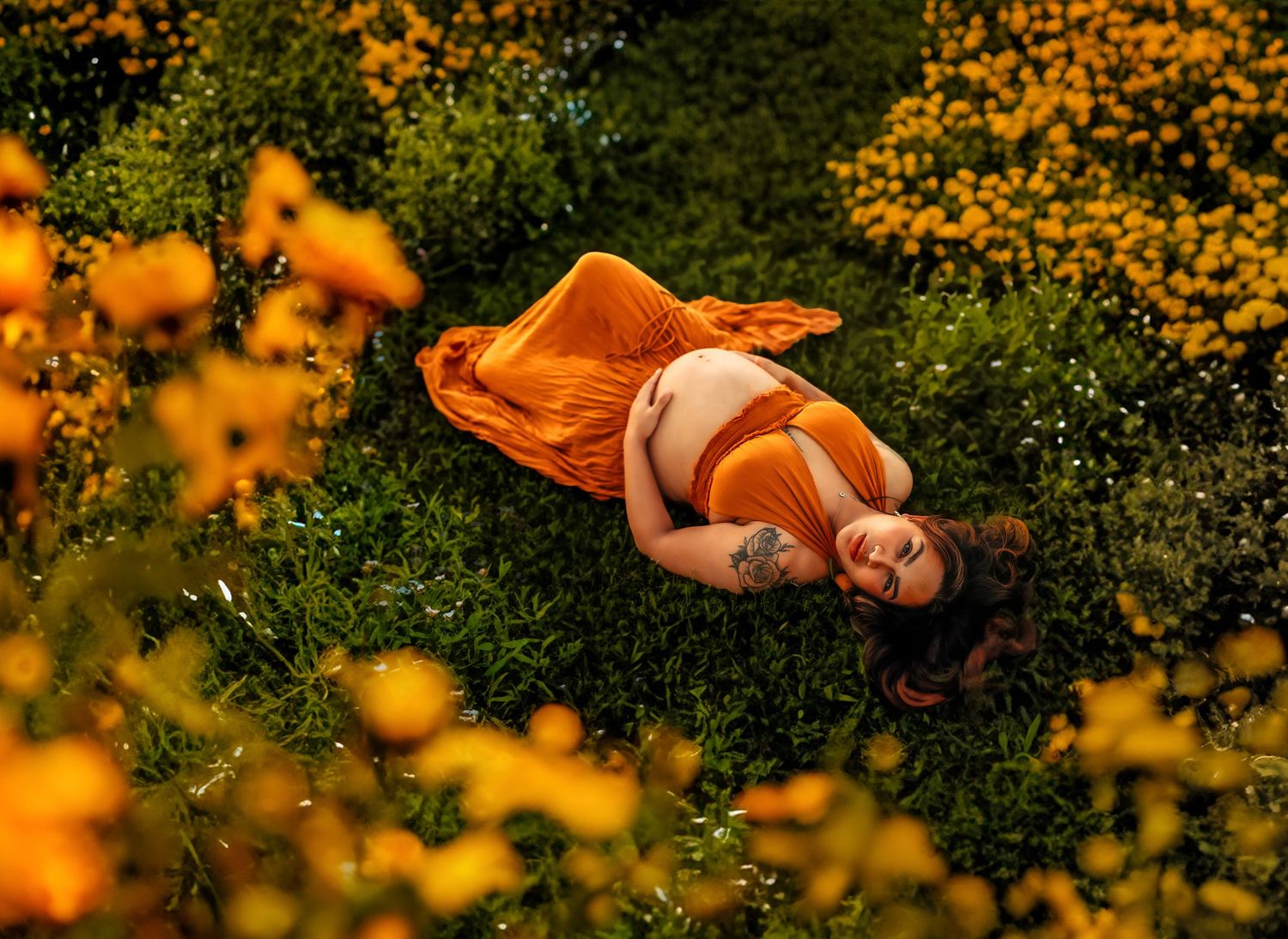 Pregnant woman in orange dress lies peacefully among yellow wildflowers outdoors.