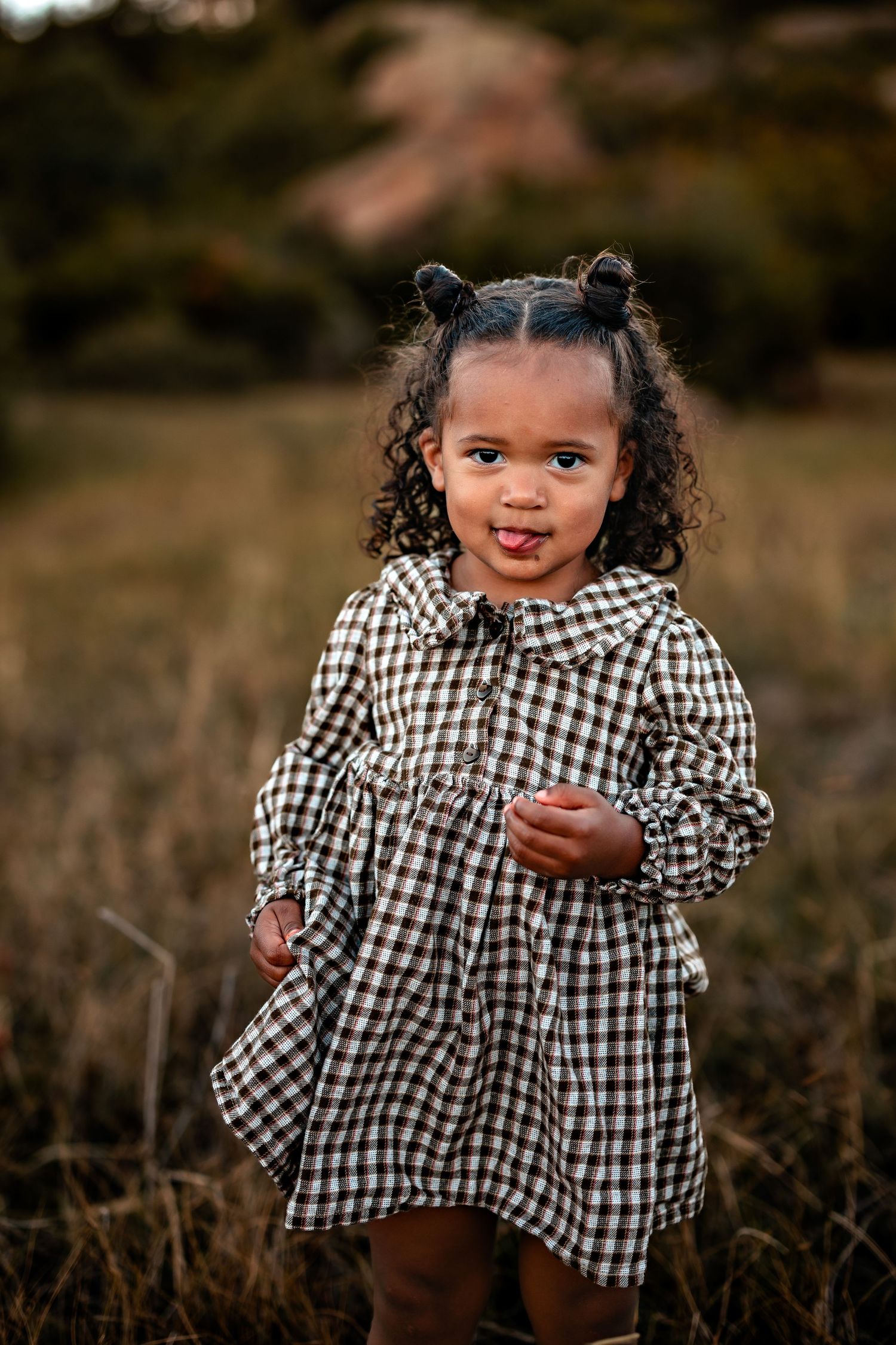 Smiling toddler with curly hair in two buns wears a brown plaid dress and sticks out her tongue while standing in a grassy field.