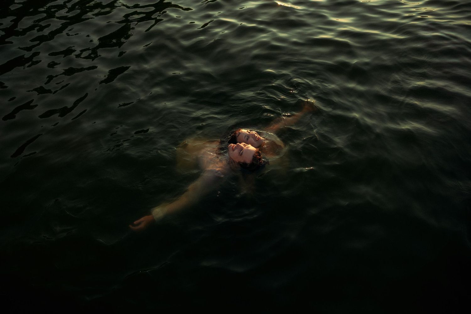 Person floating on their back in dark, rippling water during twilight, illuminated by soft warm light.