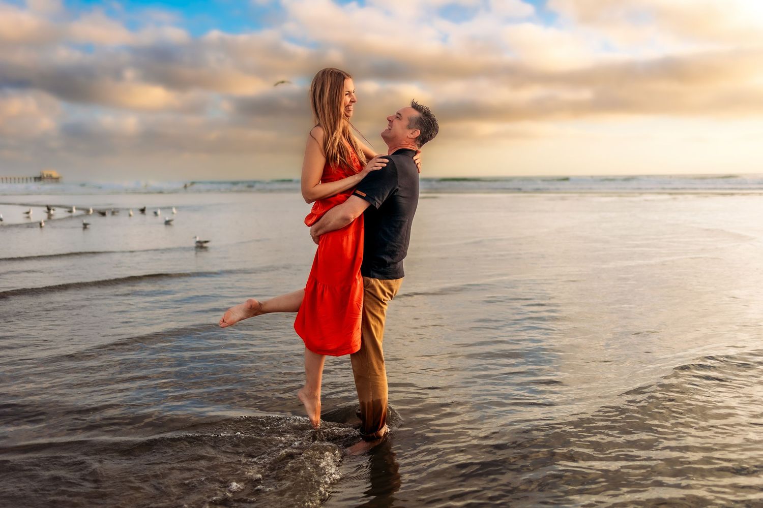 Couple embracing at sunset on a beach, woman in red dress lifted by man in casual attire, waves and seagulls in background.