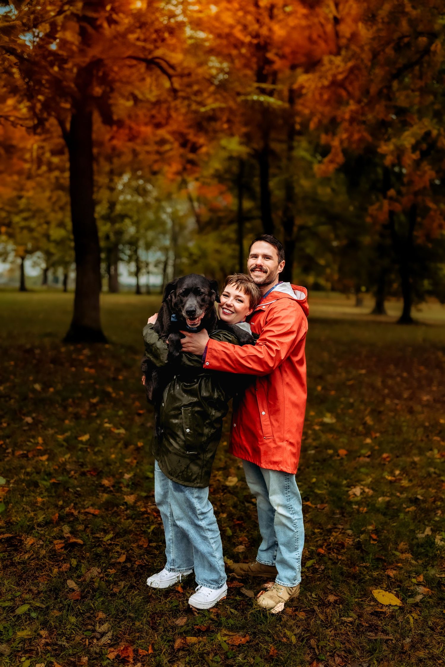 Couple hugging their black dog in an autumn park with vibrant orange foliage.