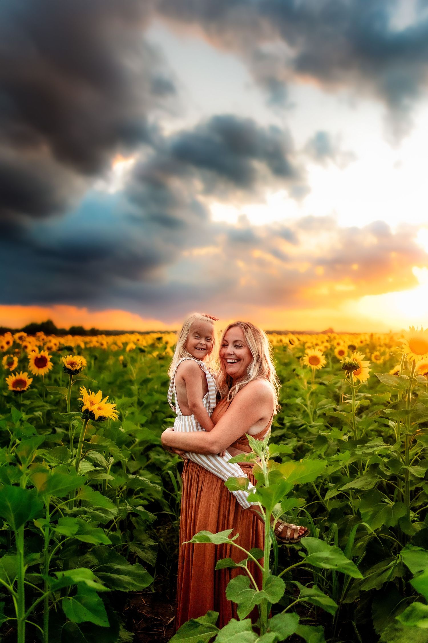 Mother holding young daughter in a sunflower field at golden sunset with dramatic storm clouds overhead.