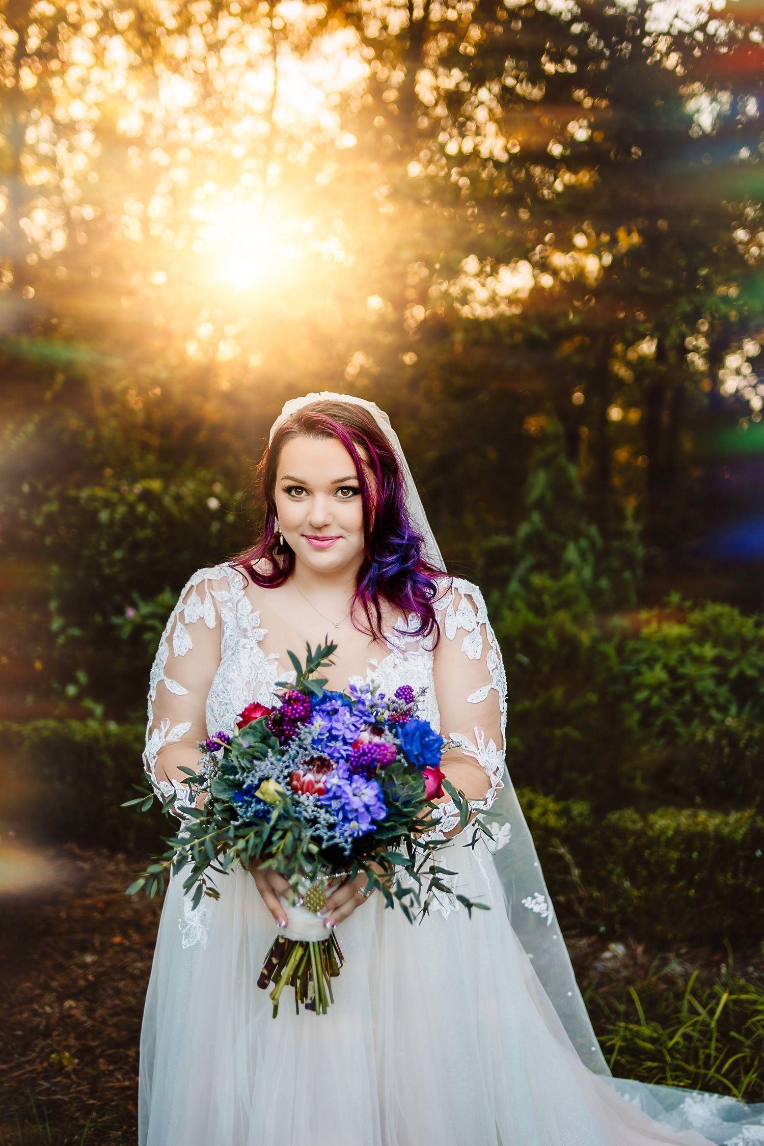 Bride in lace wedding gown holding colorful bouquet with purple hair, standing in golden sunset light outdoors.