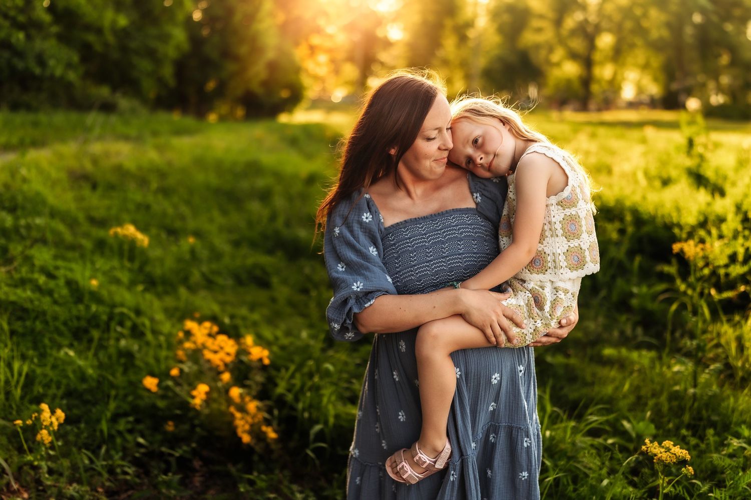 A mother holds her young daughter in a sunlit green field with yellow flowers during golden hour.