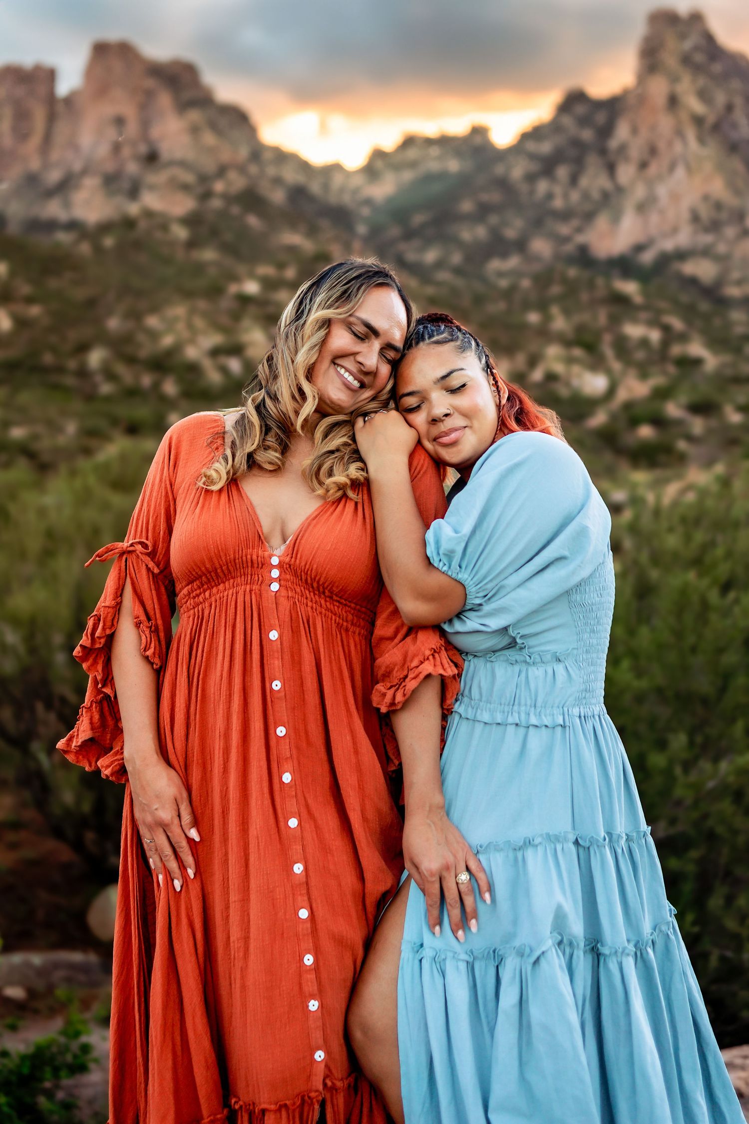 Two women in colorful maxi dresses embrace warmly at sunset with rocky mountain landscape in background.
