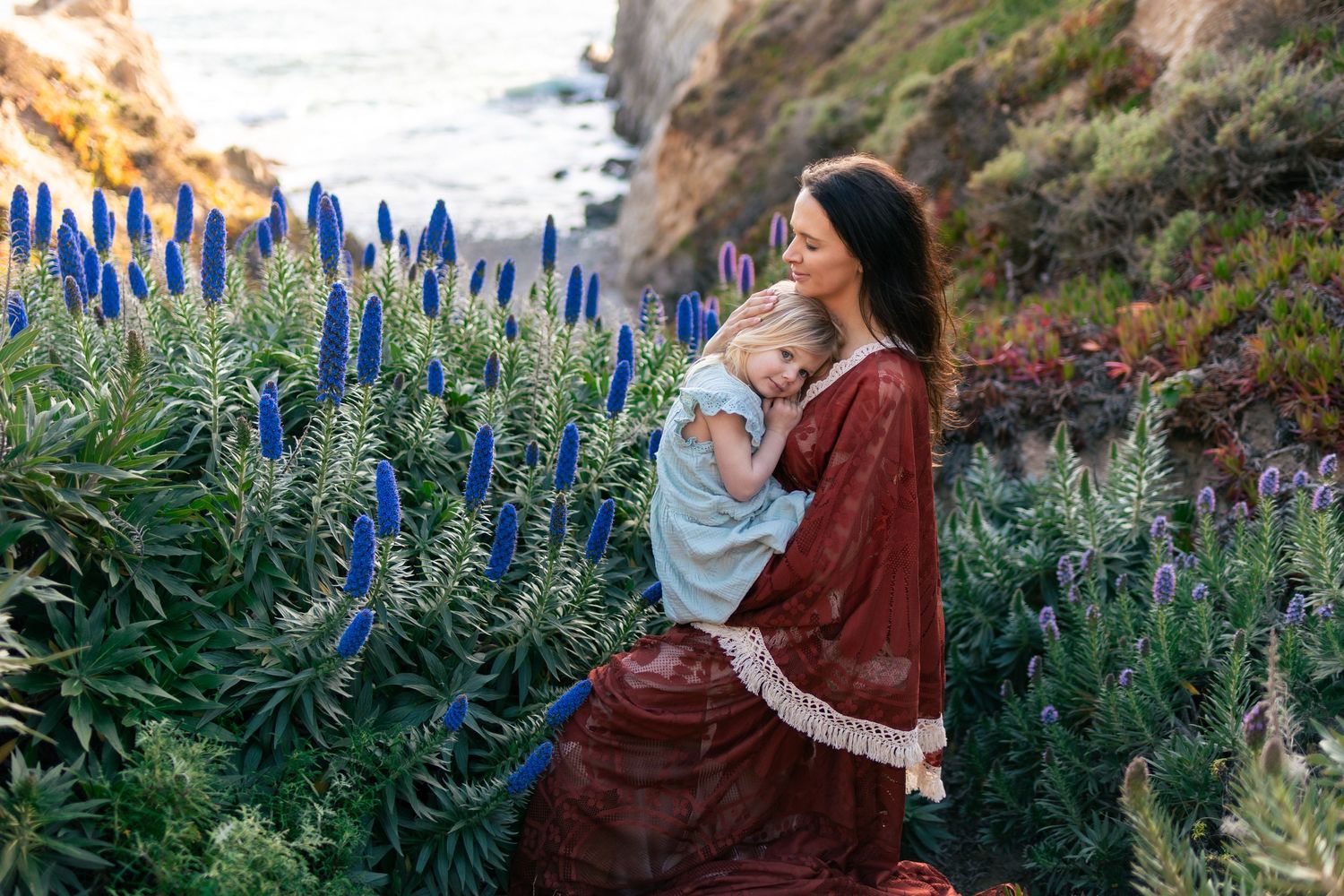 A mother in a red dress tenderly holds her child among blue wildflowers on a scenic coastal cliffside.