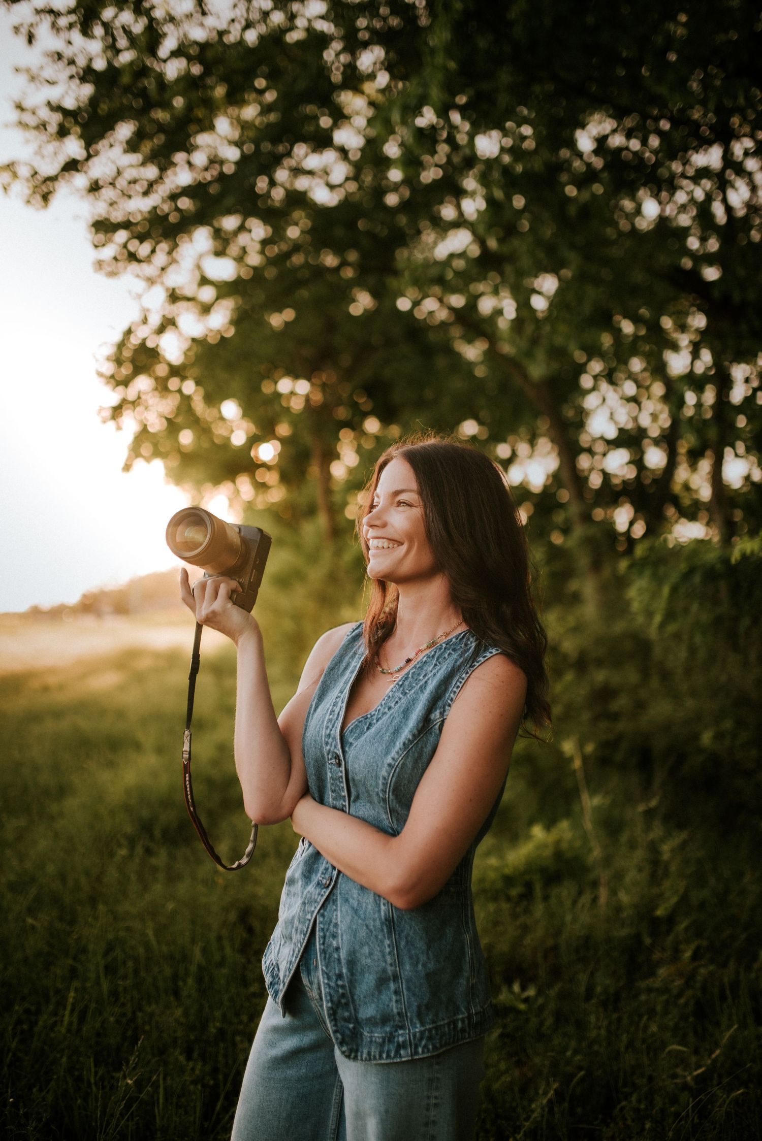 Smiling brunette woman holding camera in denim vest at golden hour in lush green outdoor setting.