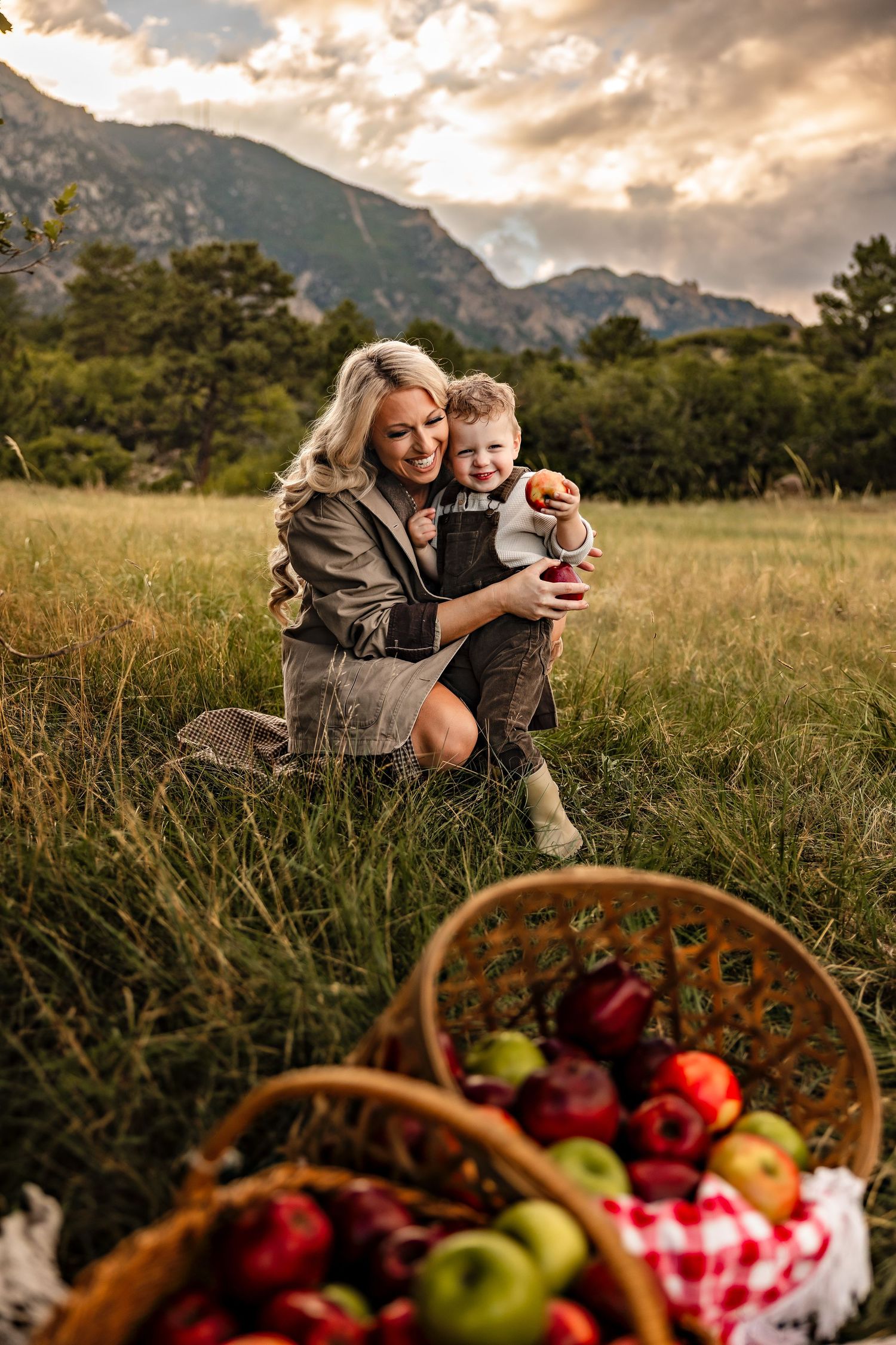 Smiling mother kneeling in a grassy mountain meadow holding her toddler, with baskets of red and green apples in the foreground.