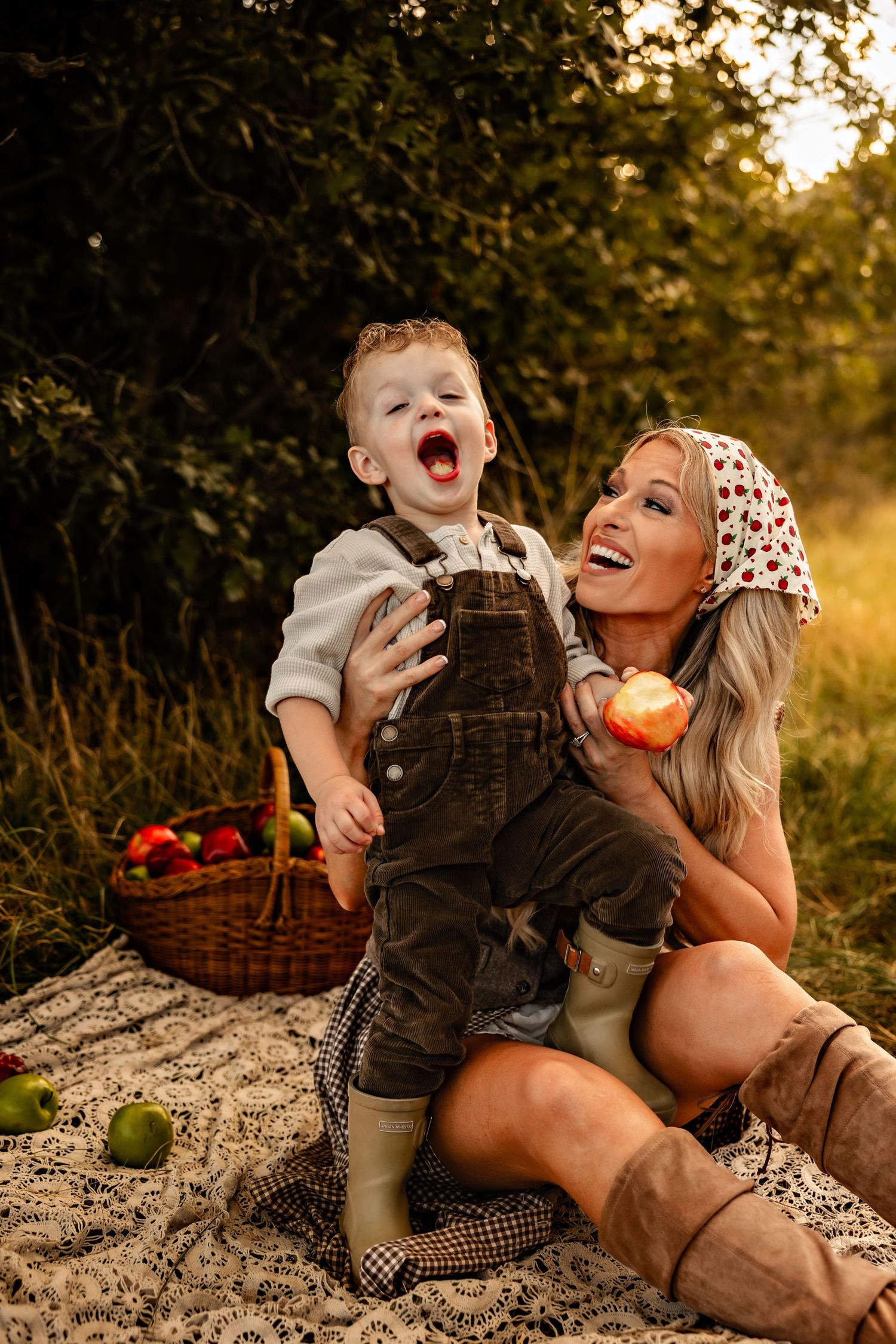Smiling mother in a polka dot headscarf holds a laughing toddler in overalls on a picnic blanket with apples in a basket outdoors.
