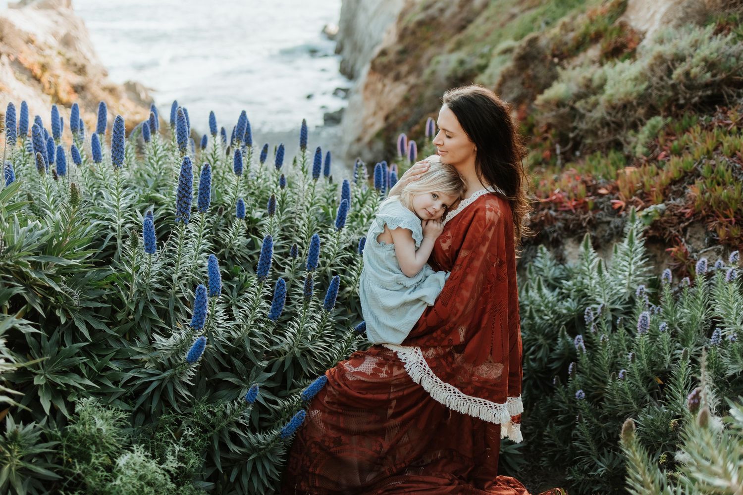 Woman in red dress holds baby among blue Pride of Madeira flowers near coastal cliffs.