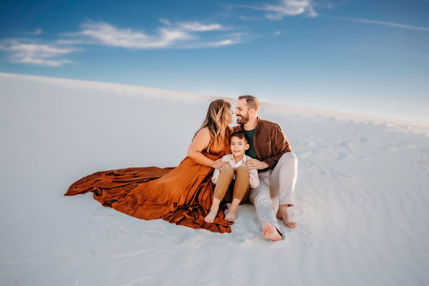 Family of three sitting on white sand dunes, woman in flowing rust gown, man kissing her cheek, child between them.