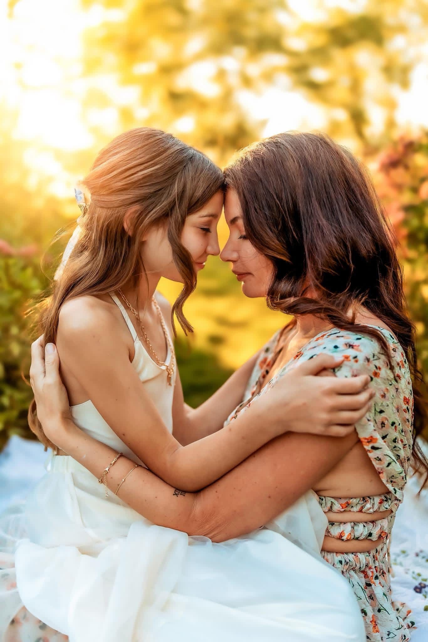 Two young women in summer dresses embrace tenderly outdoors, touching foreheads in golden sunset light.