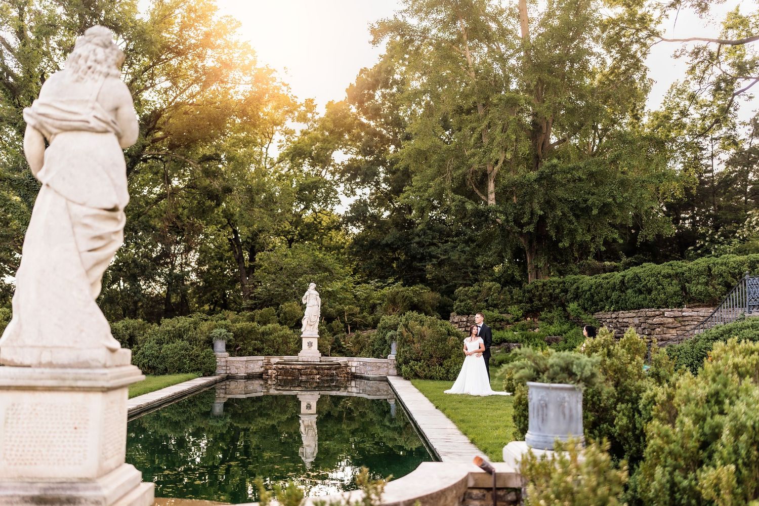 Bride and groom pose near a reflecting pool surrounded by classical statues and lush gardens at golden hour.