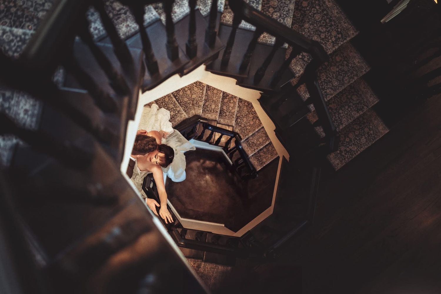 Aerial view of a woman lying at the bottom of a dark wooden spiral staircase, shot from above.