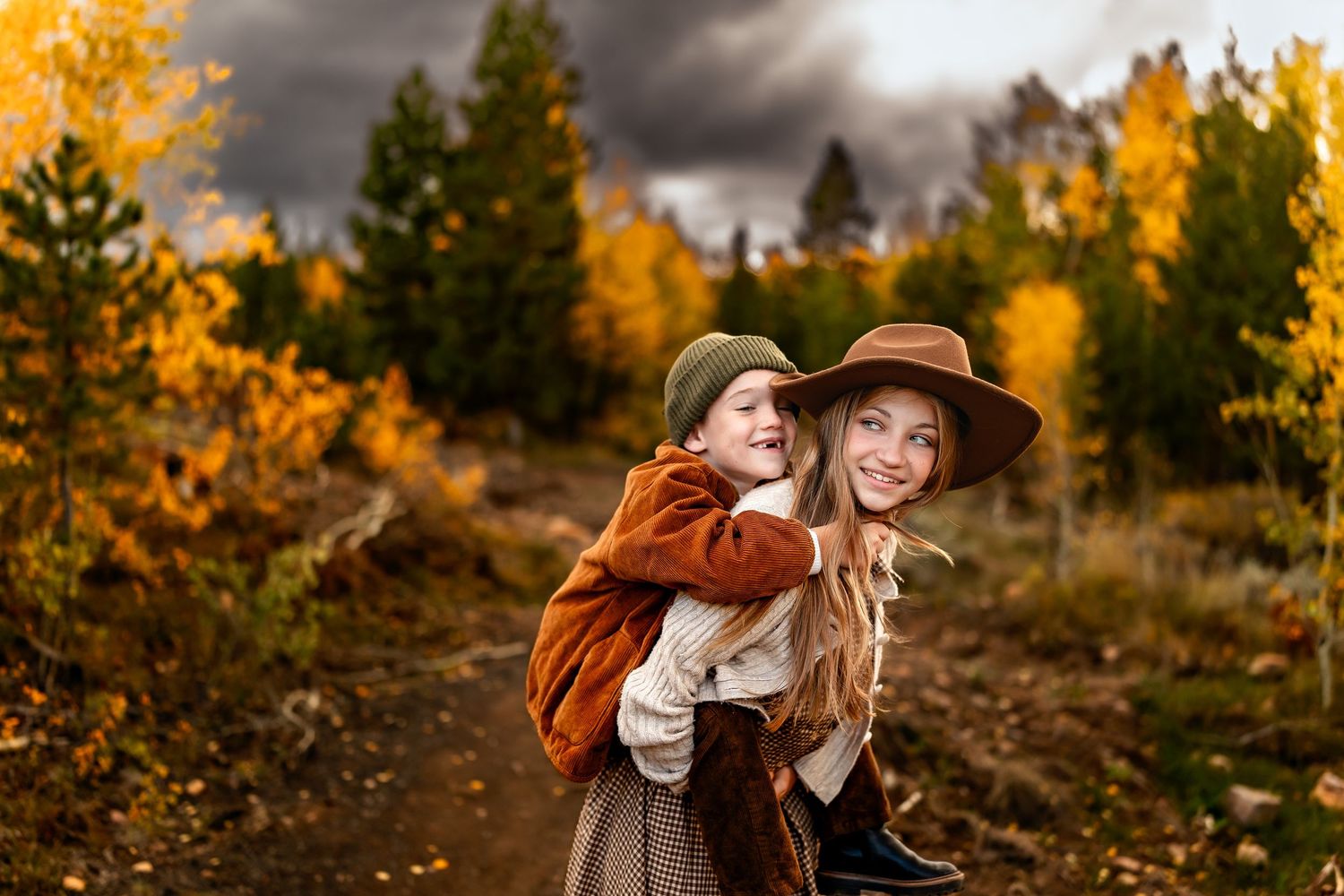 Two children laughing in autumn forest, girl giving piggyback ride, wearing fall hats, colorful foliage background.