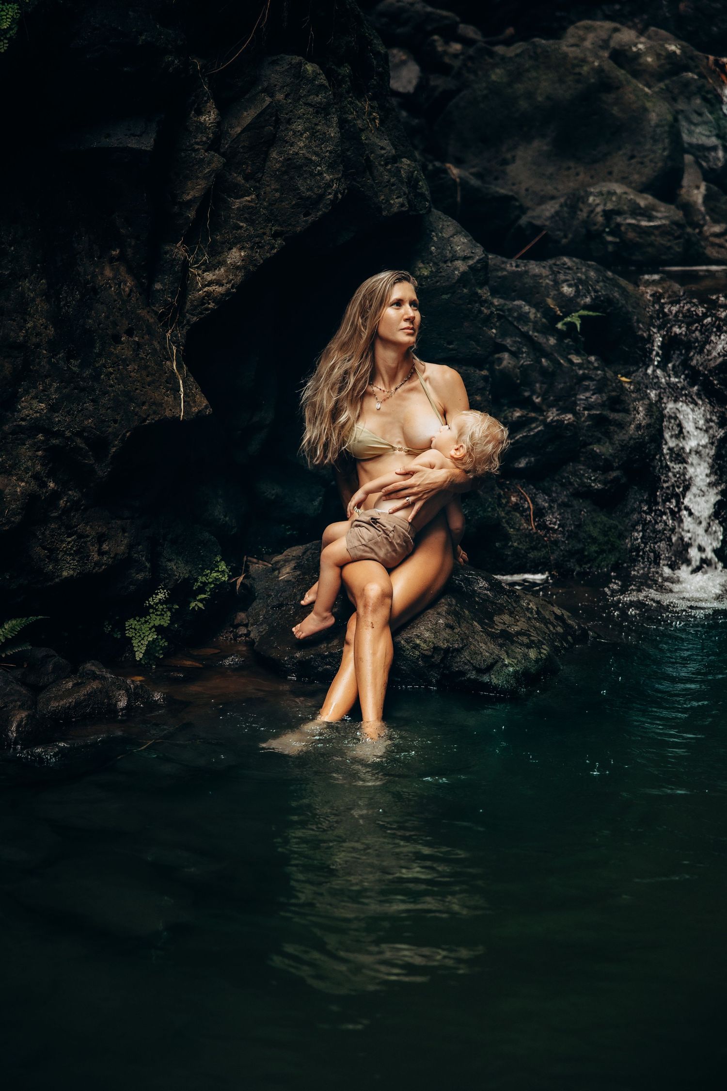 A woman in a bikini sits on rocks by a waterfall, cradling a baby while her feet rest in the water.