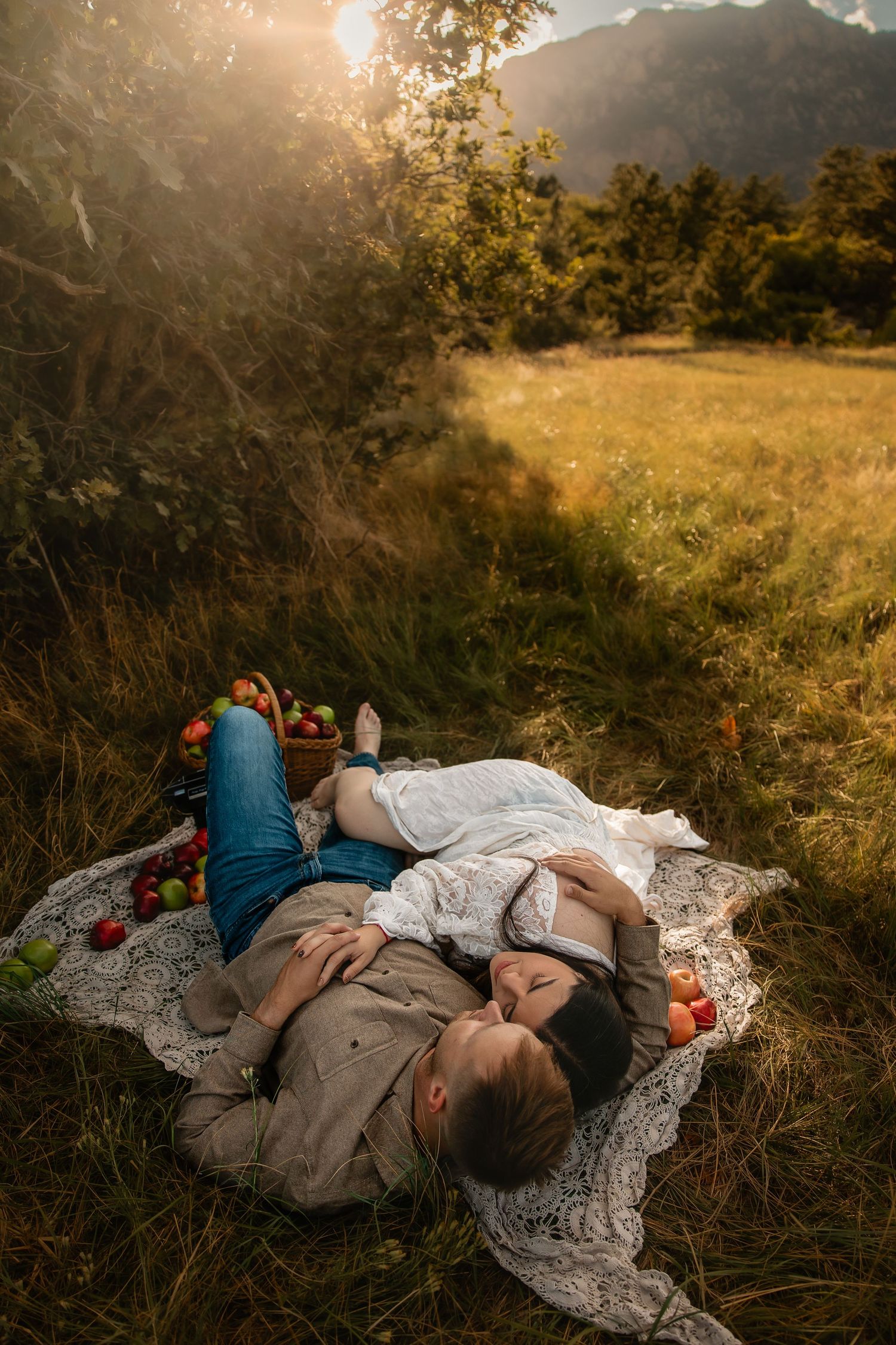 Couple lying on lace blanket during golden hour picnic in mountain meadow with apples and warm sunlight.
