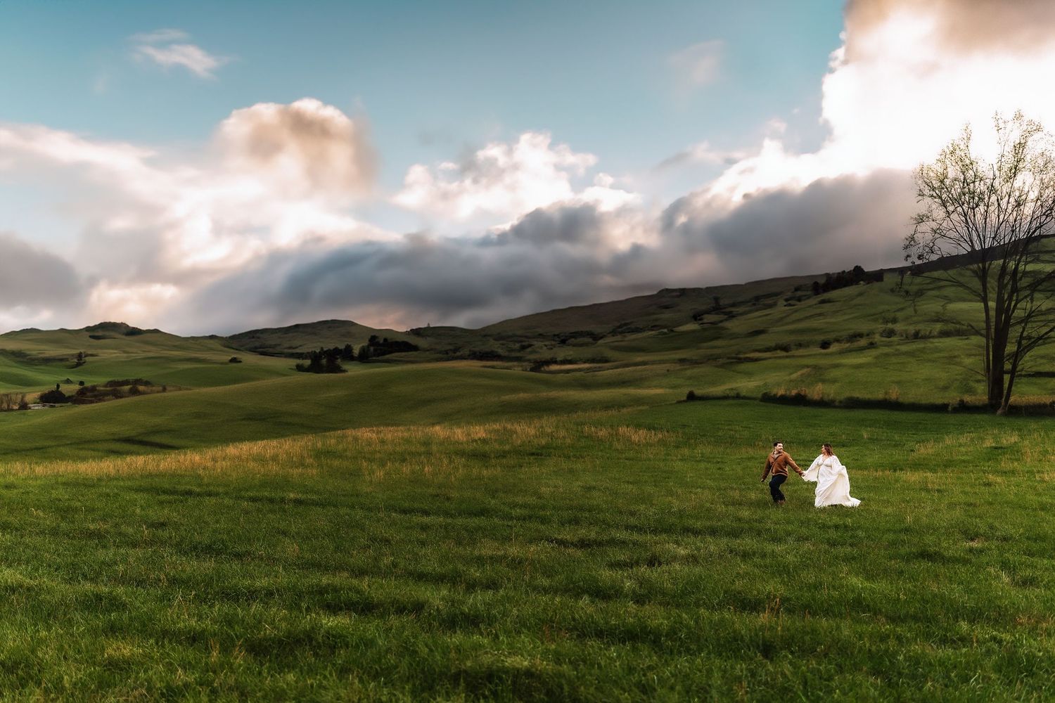 Bride and groom walking through lush green rolling hills under dramatic cloudy sky at golden hour.