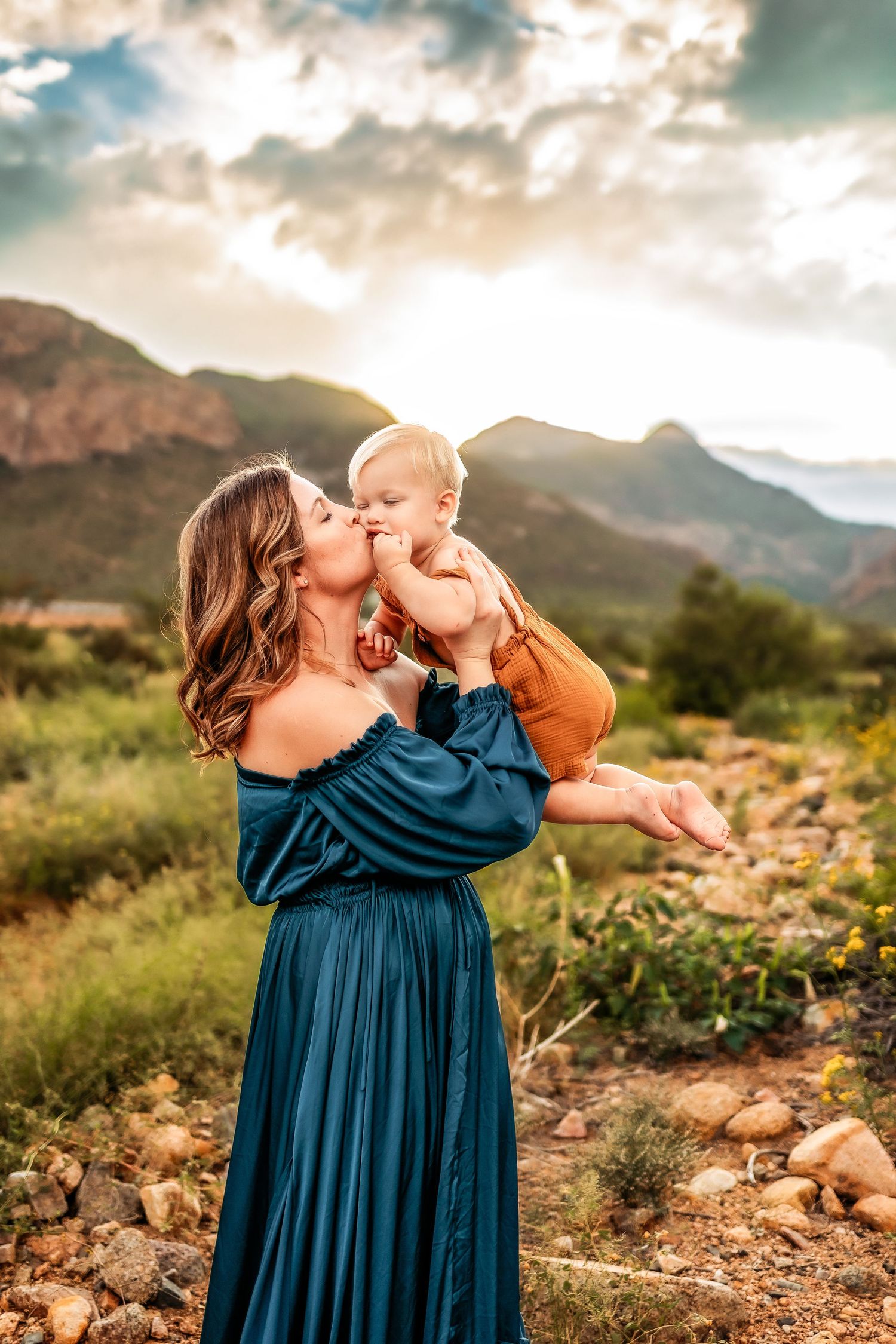 A mother in a teal dress kisses her baby outdoors with mountains and a dramatic sky in the background.