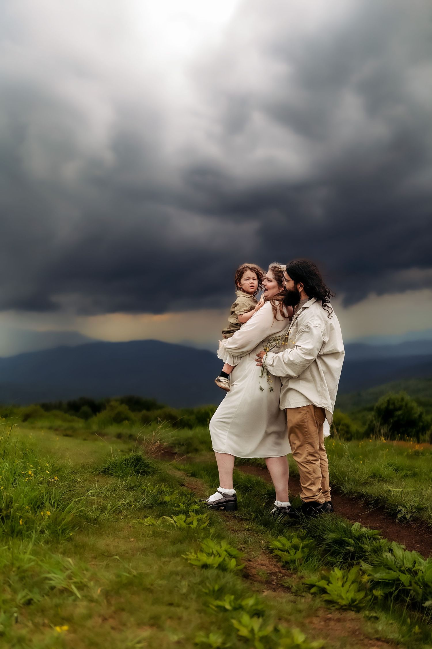 Family of three standing on a grassy mountain path under dramatic stormy skies, mother holding child.