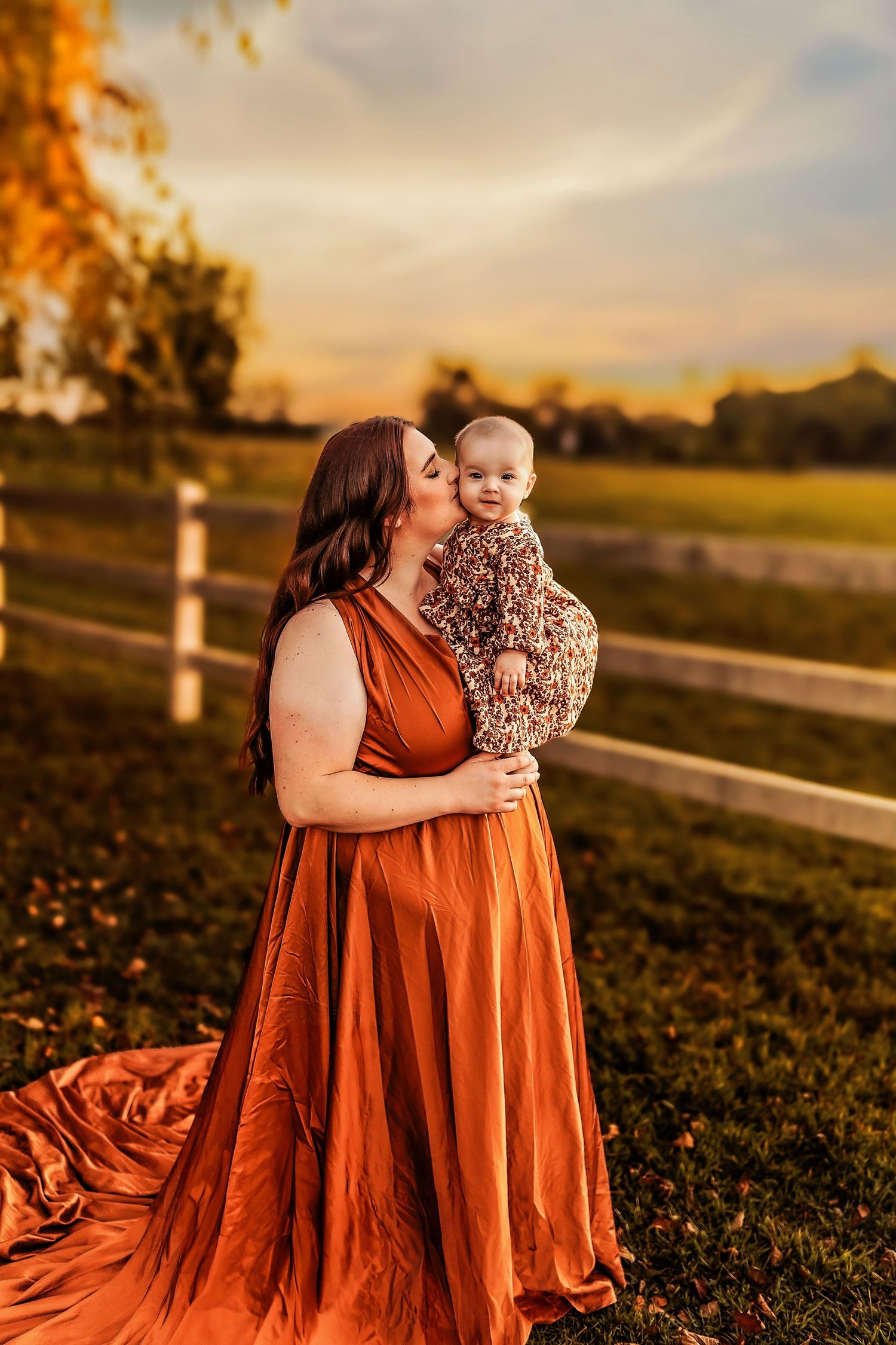 Mother in orange gown kissing baby at golden hour sunset near white fence in autumn field.