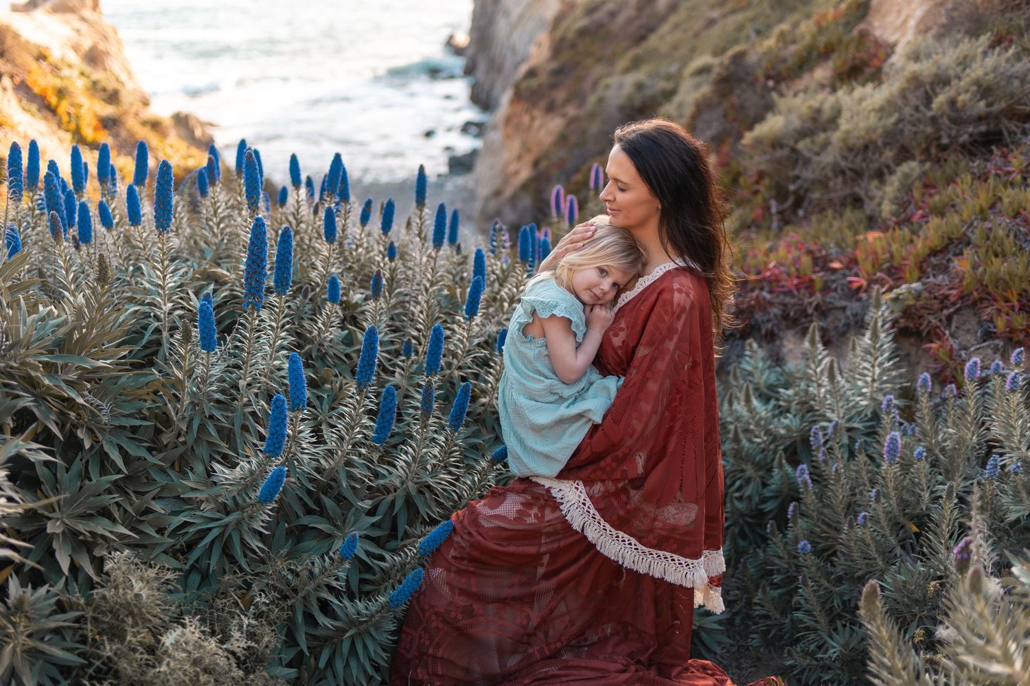 Woman in red dress holds a child among blue wildflowers on a coastal cliffside at golden hour.