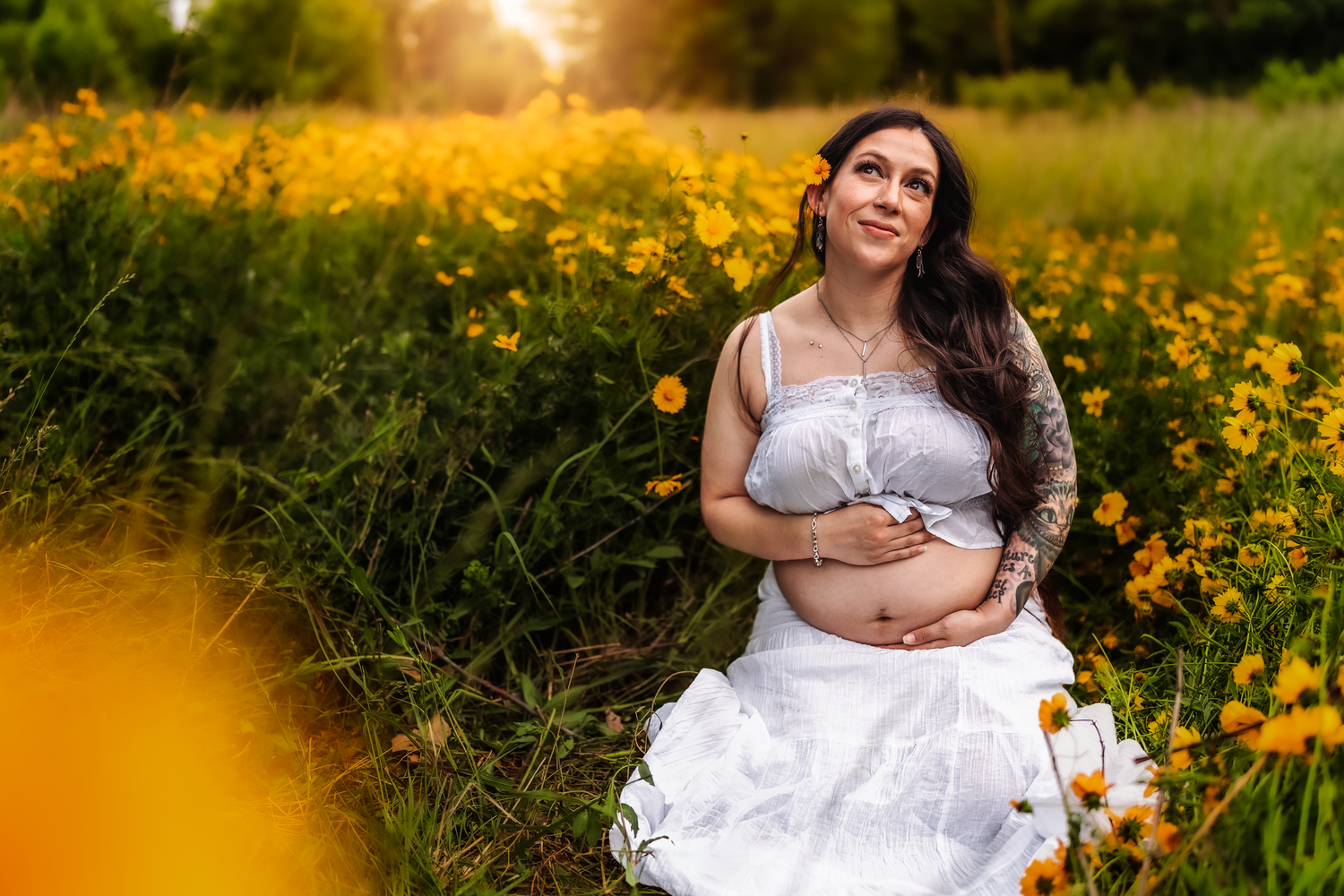 Pregnant woman in white dress sitting among yellow wildflowers at sunset, cradling her baby bump.