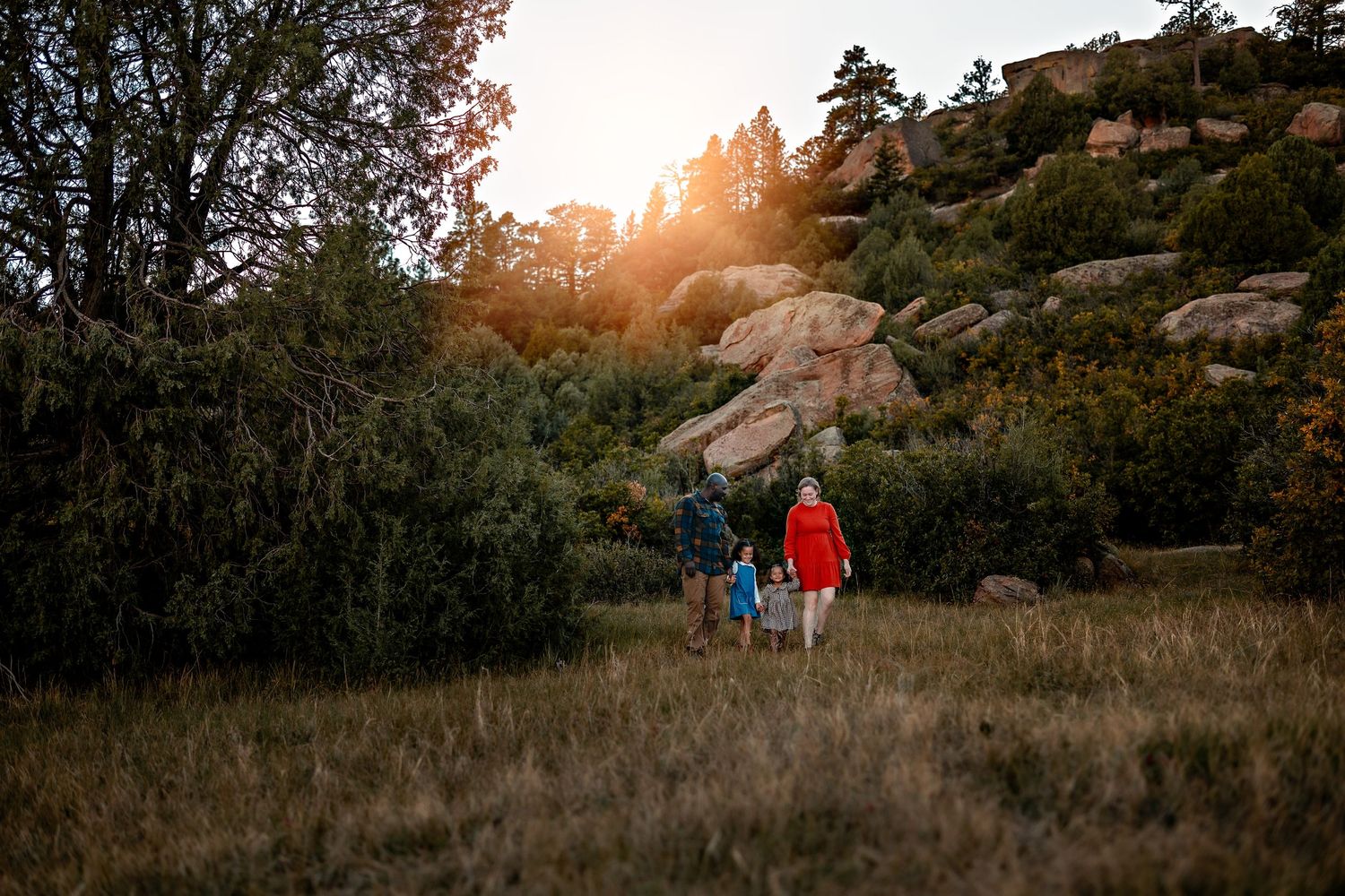 Family of four in colorful jackets walks through a grassy meadow toward rocky boulders and pine trees at golden hour sunset.