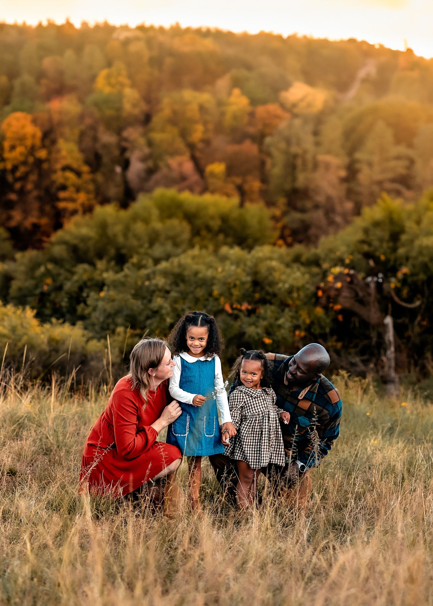 Family of four with two young girls in matching outfits smiling together in a golden grassy field with colorful autumn trees behind them.