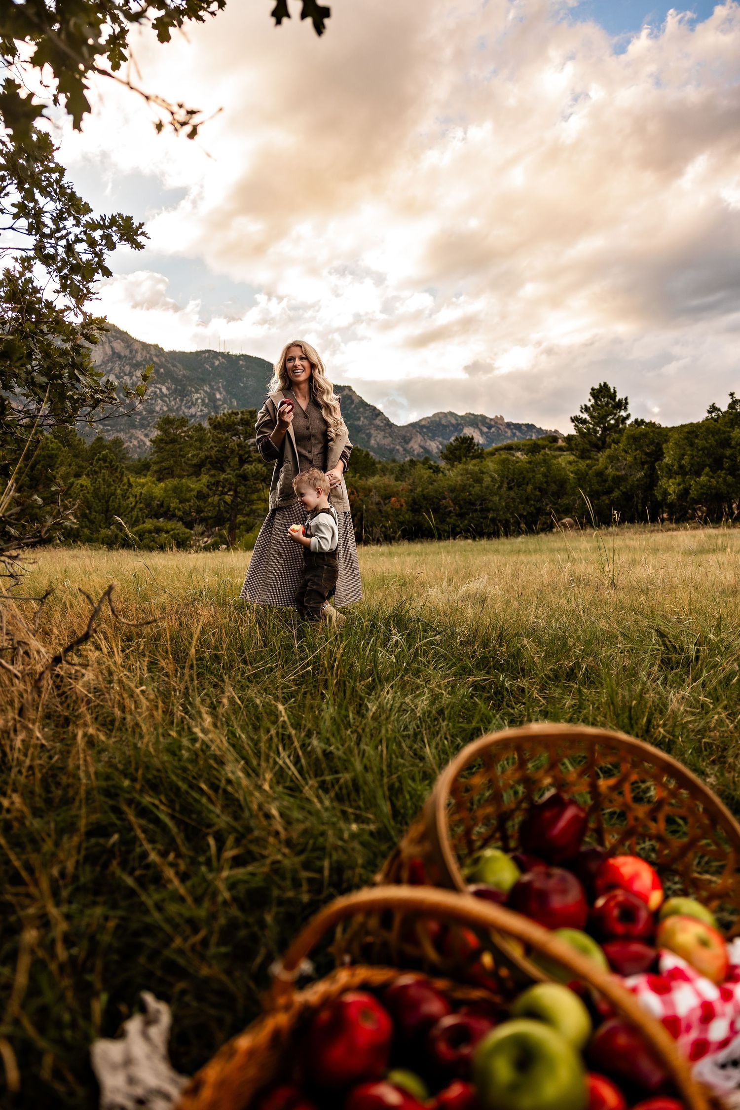 Woman and toddler stand in a grassy meadow with mountains behind them, while baskets of fresh red and green apples sit in the foreground.