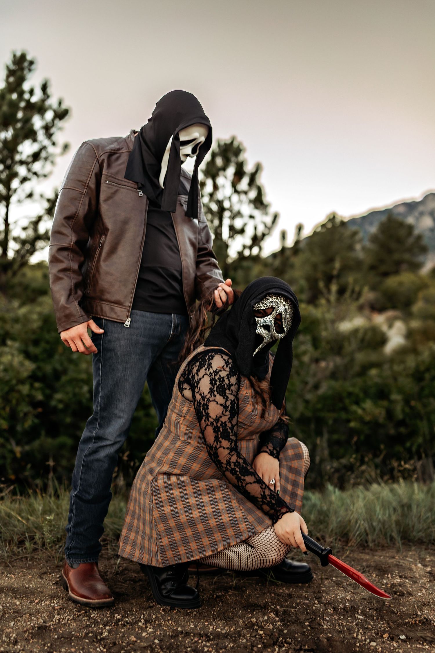 Two people in Ghostface masks and black hoods pose outdoors, one standing in a leather jacket, the other crouching with a red knife.