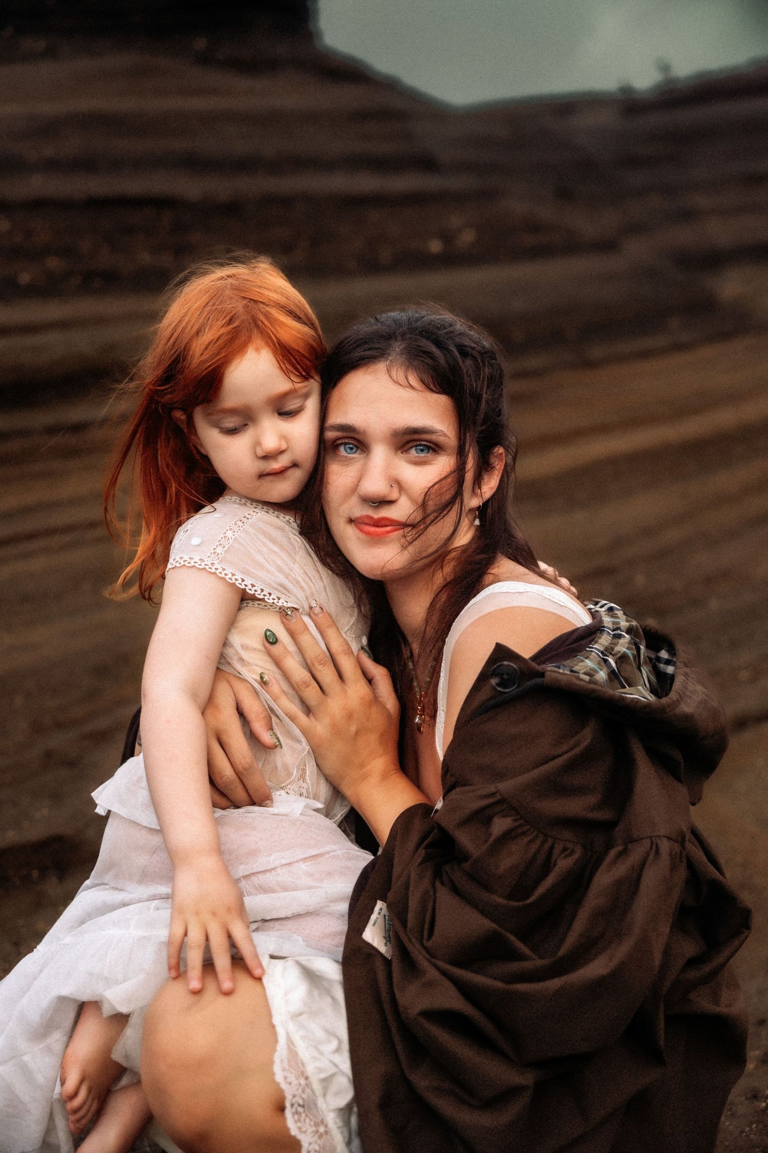 A dark-haired woman in brown dress tenderly embraces a red-haired little girl in white dress outdoors.