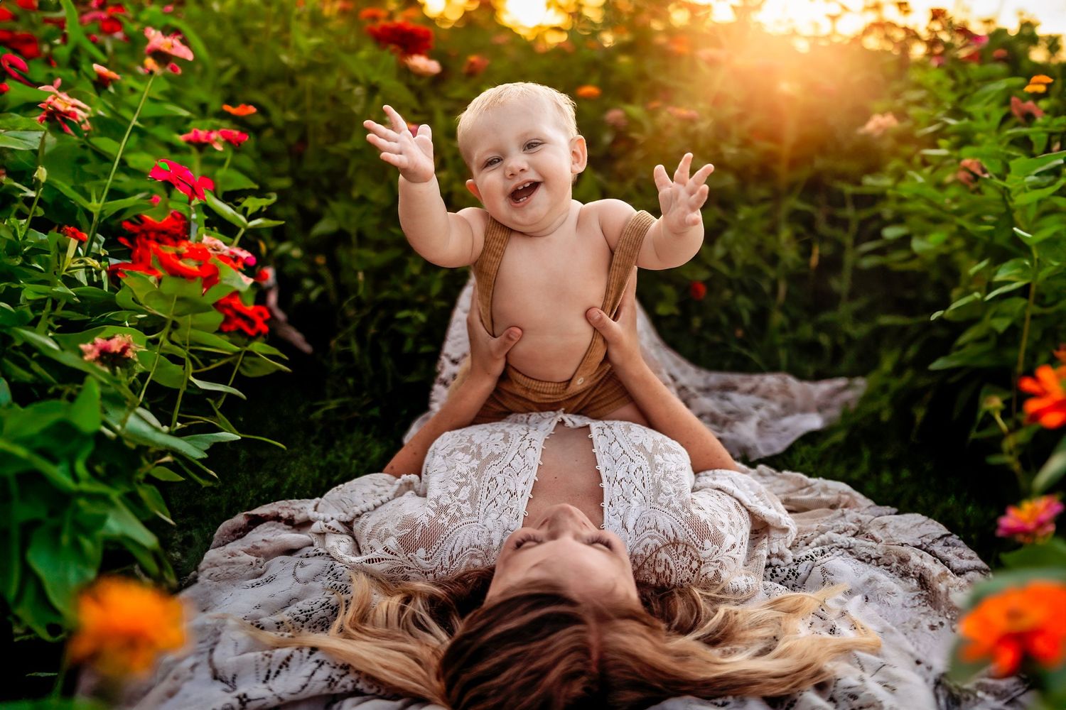 A joyful baby laughs while being lifted by a mother lying in a lush garden with red and orange flowers.