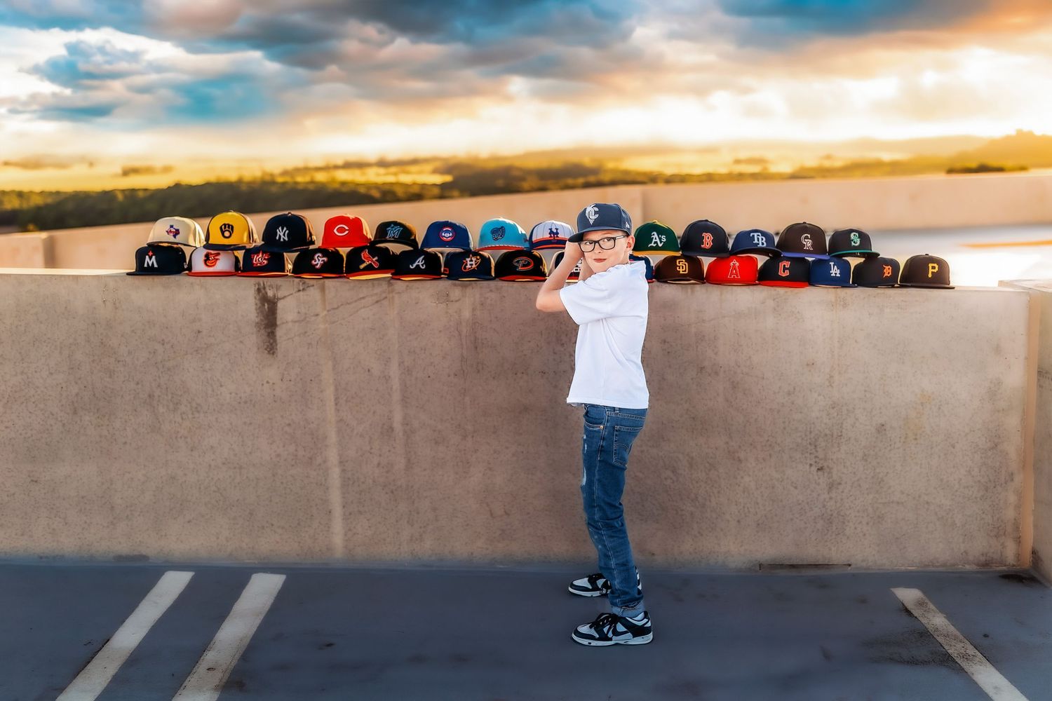 Young boy posing with a baseball bat surrounded by MLB caps displayed on a concrete wall at sunset.