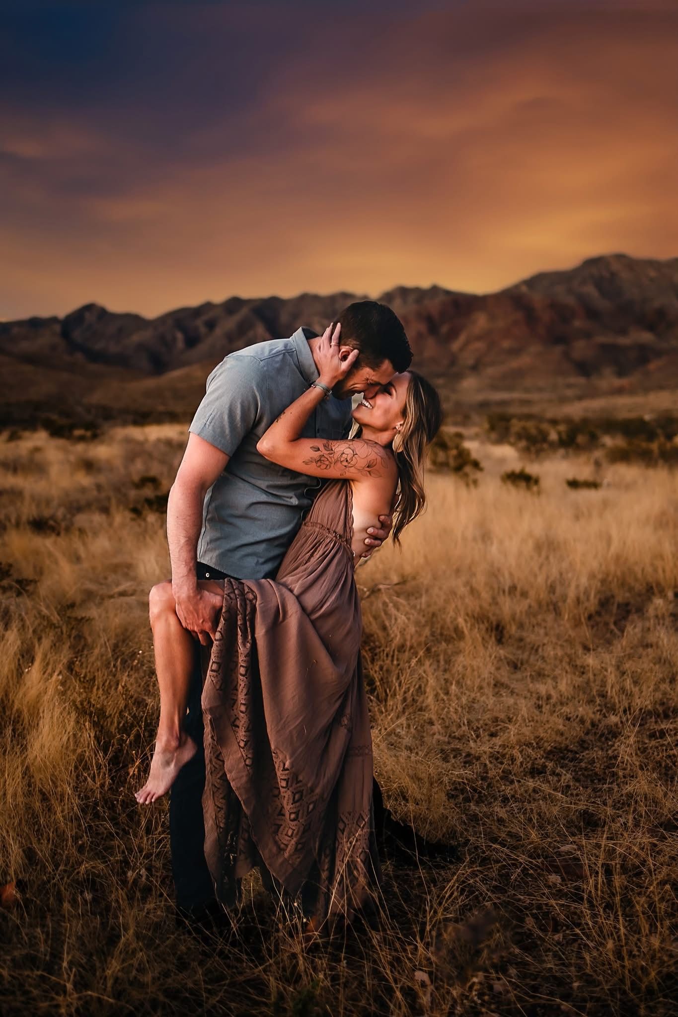 A couple shares a romantic kiss at sunset in a desert landscape with mountains in the background.