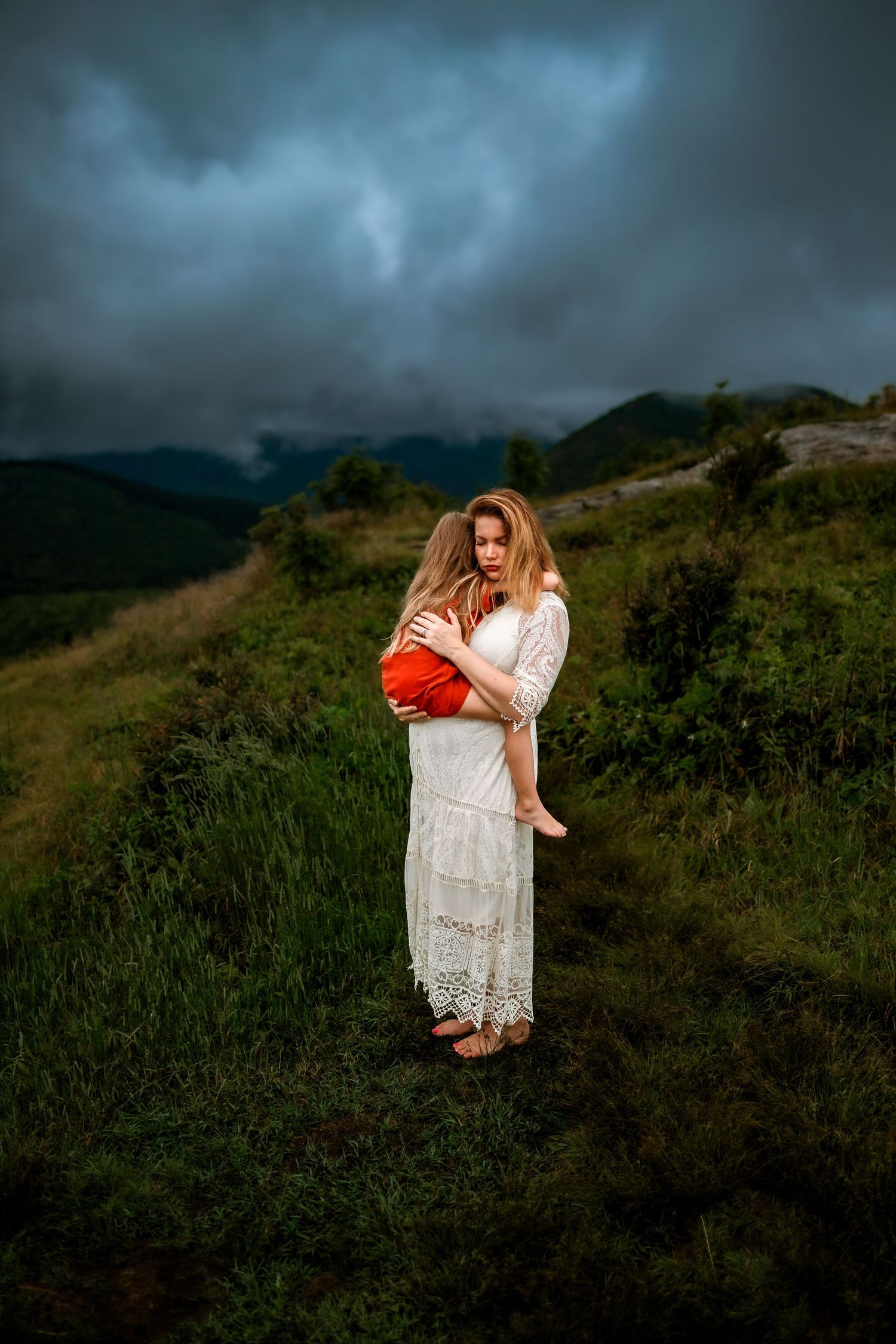 Mother in white lace dress holds young child on a green hillside under dramatic stormy skies.