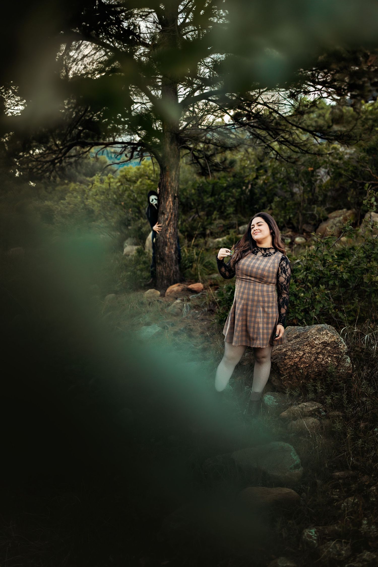 Smiling woman in a plaid dress stands on a rocky forest hillside while a figure in a ghost mask peeks from behind a pine tree.