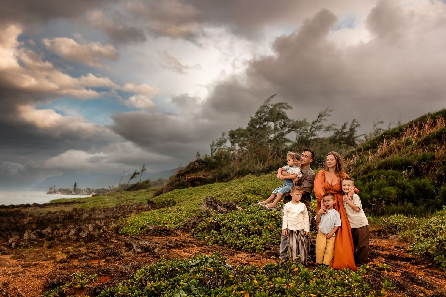 Family of five posing on a lush green coastal hillside under dramatic cloudy skies at golden hour.