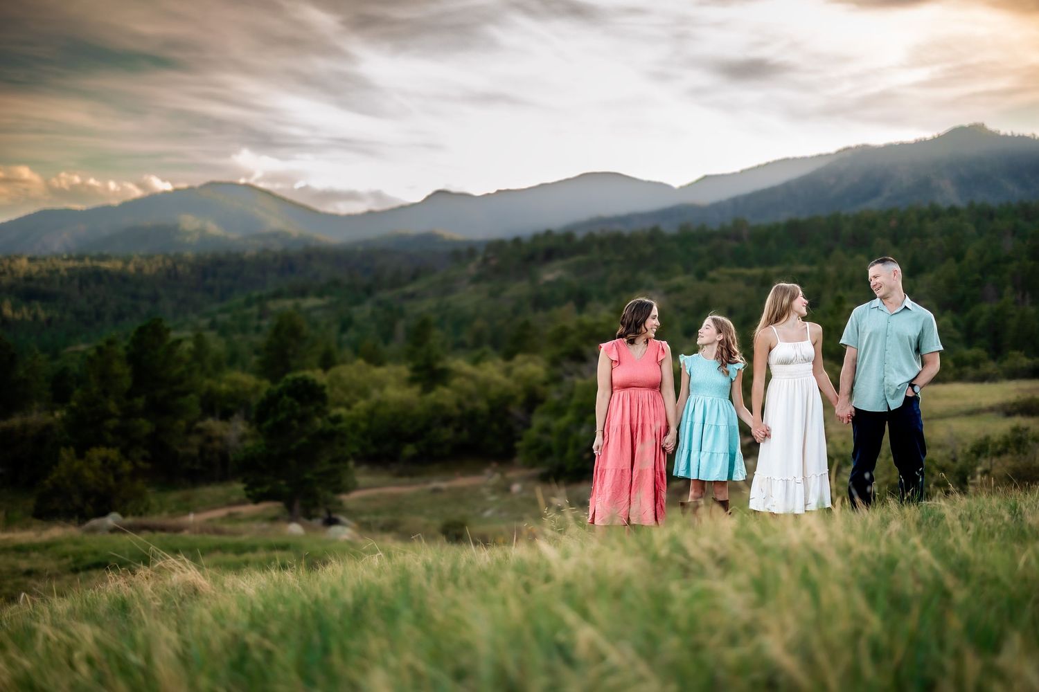 Family of four holding hands in a meadow with mountain views and dramatic sunset sky in background.
