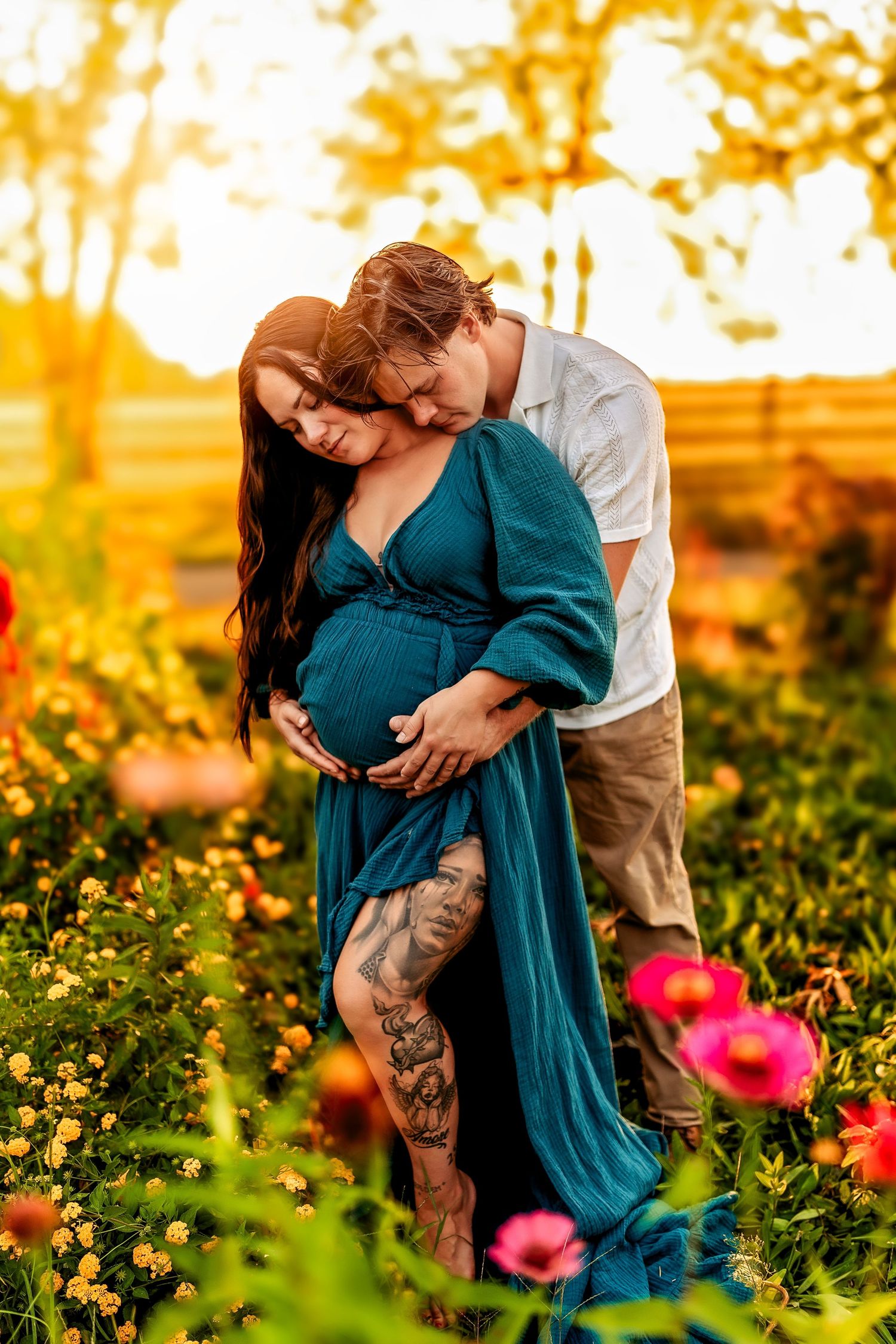 Pregnant couple embracing in colorful flower garden at golden sunset, woman in flowing teal gown.